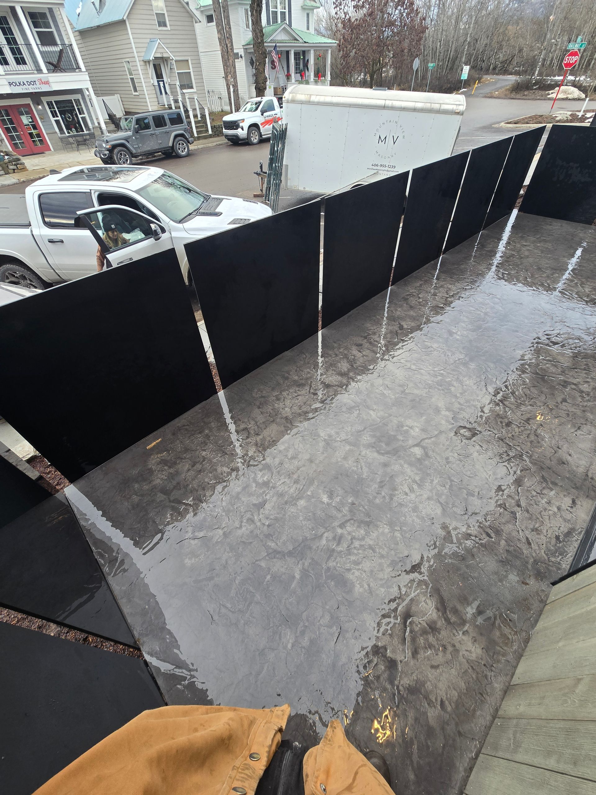 A view from above of a black temporary fencing barrier surrounding a wet, paved area with parked cars and buildings nearby.
