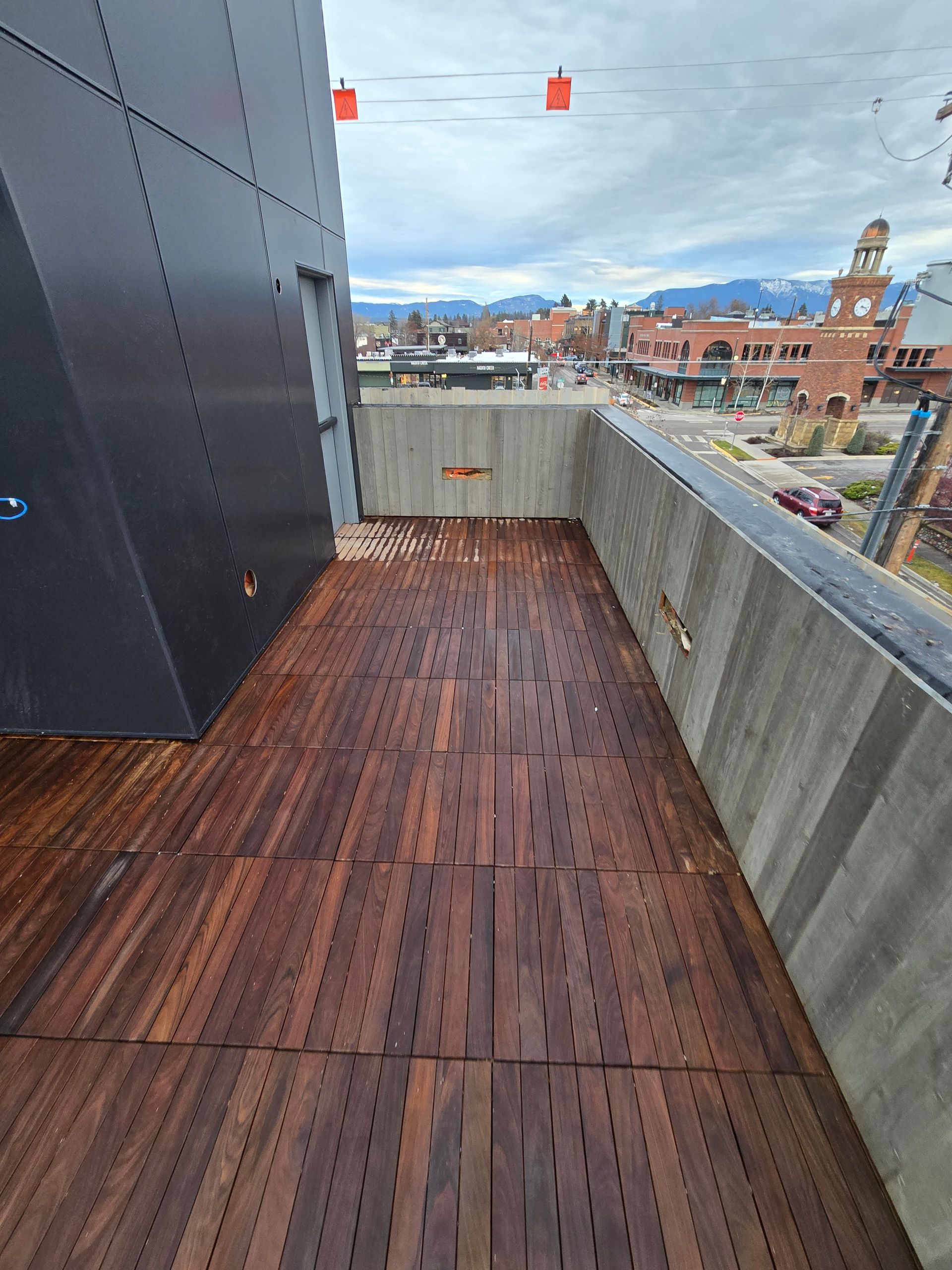 A high-angle view of a wooden rooftop deck with a grey concrete perimeter wall, overlooking a town street and mountains.