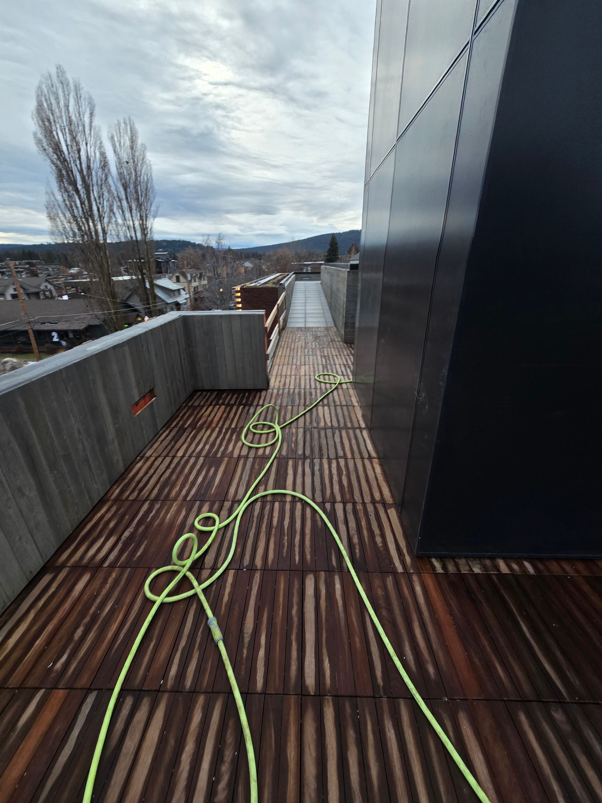 A bright green garden hose lies coiled on a wooden rooftop deck next to a tall, dark metal wall under a cloudy sky.