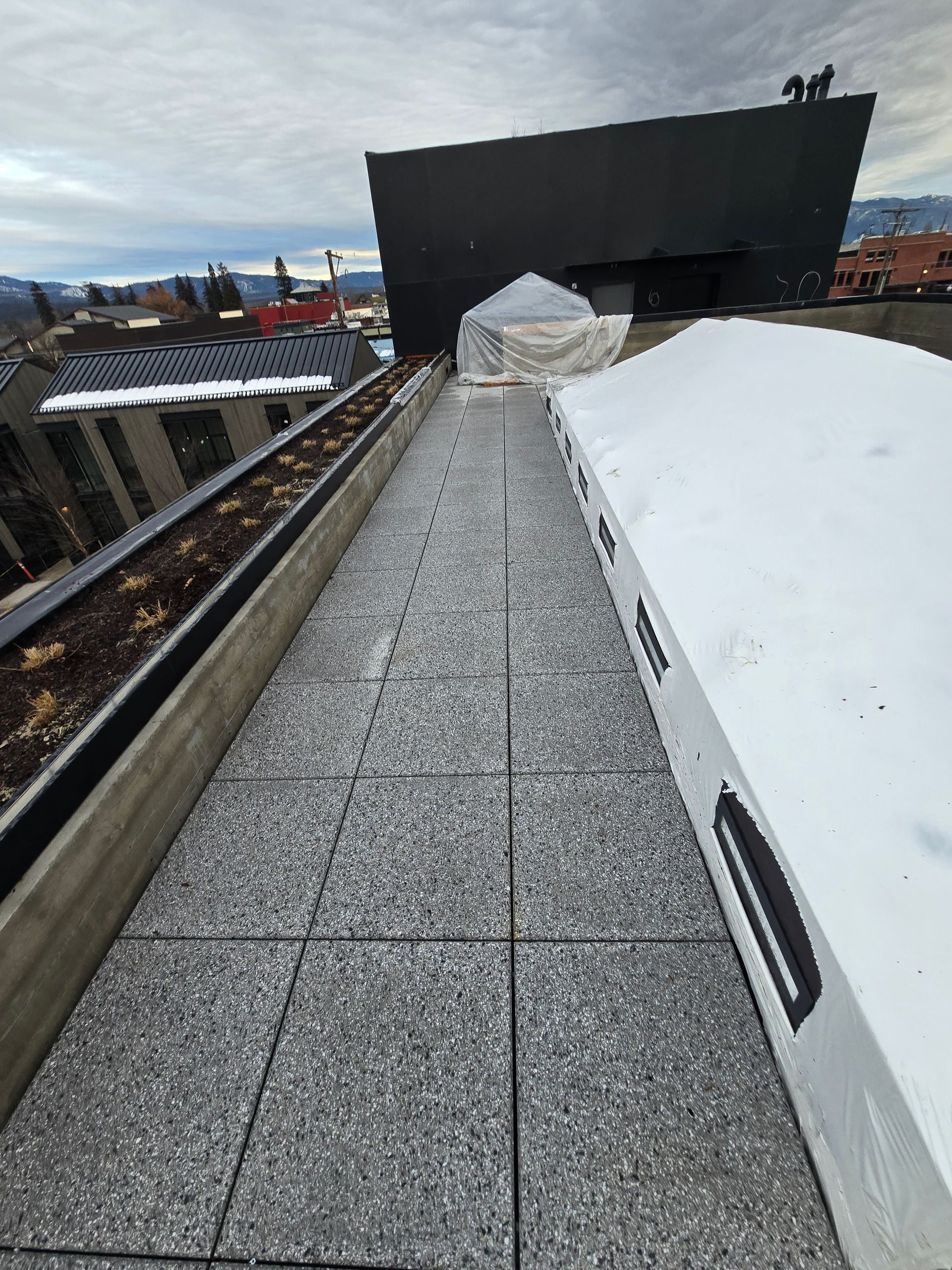 A rooftop walkway with light grey textured pavers, bordered by a planter box on the left and a snow-covered ledge on right.