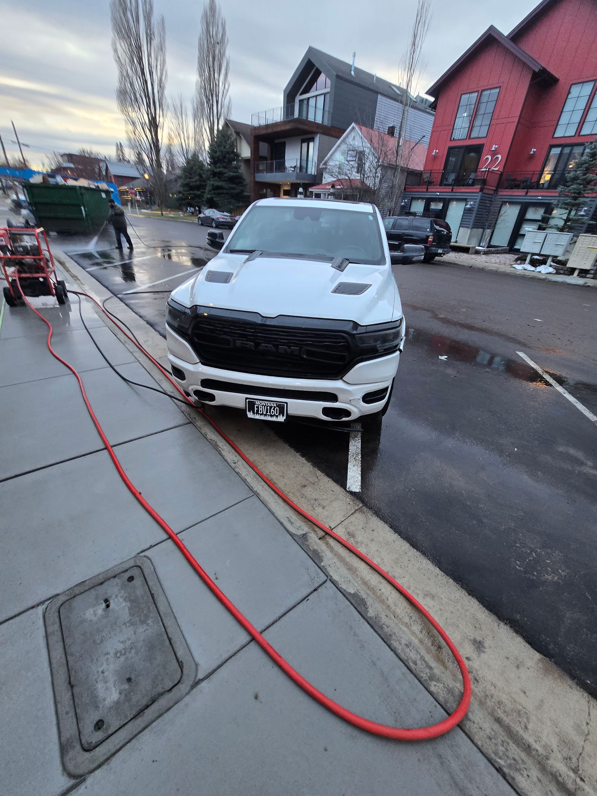 A white pickup truck parked on a wet street beside a red building, with a red hose stretching across the sidewalk.