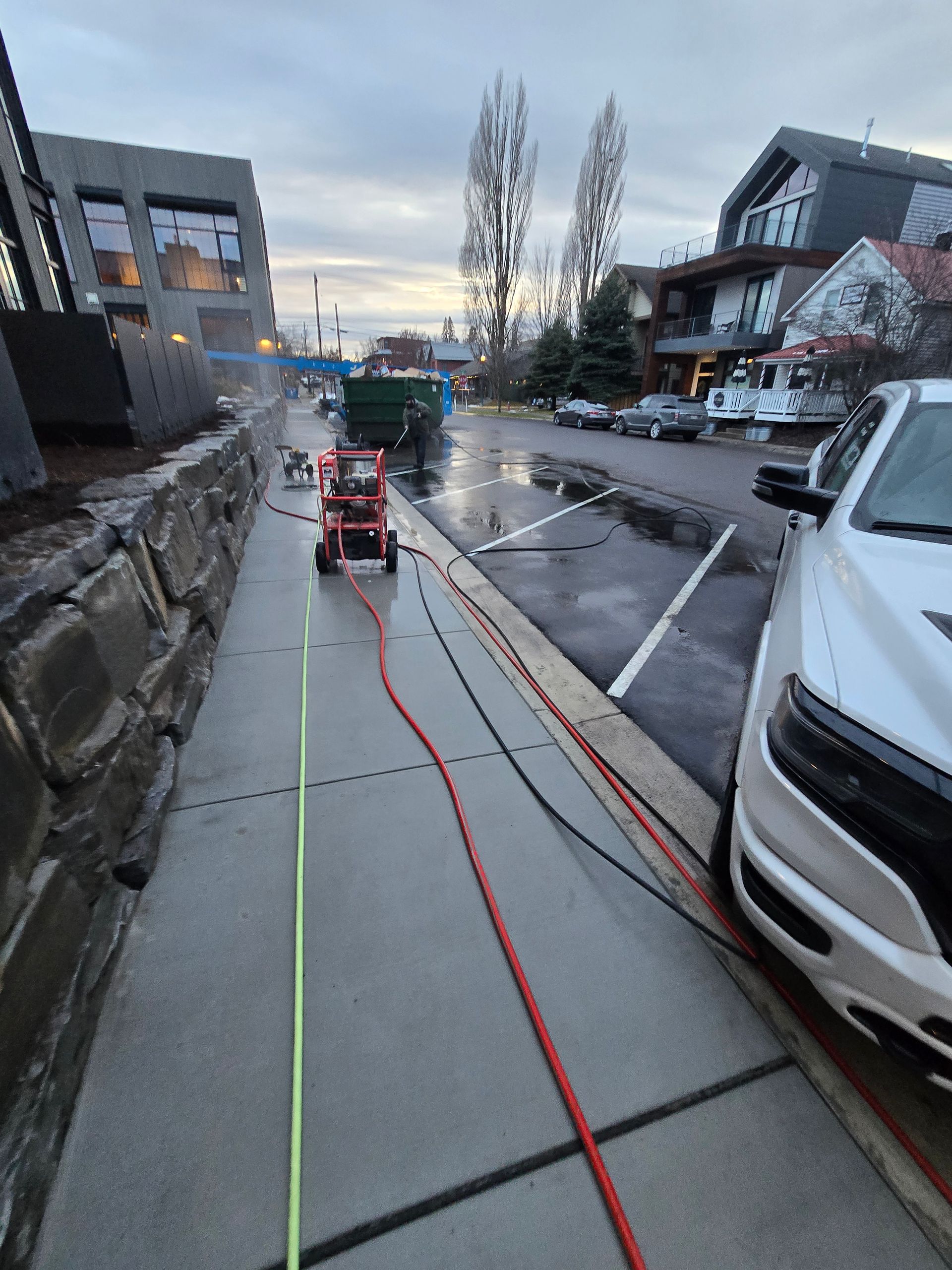 A sidewalk power washing machine with hoses extending along a walkway near parked cars and buildings at dusk.