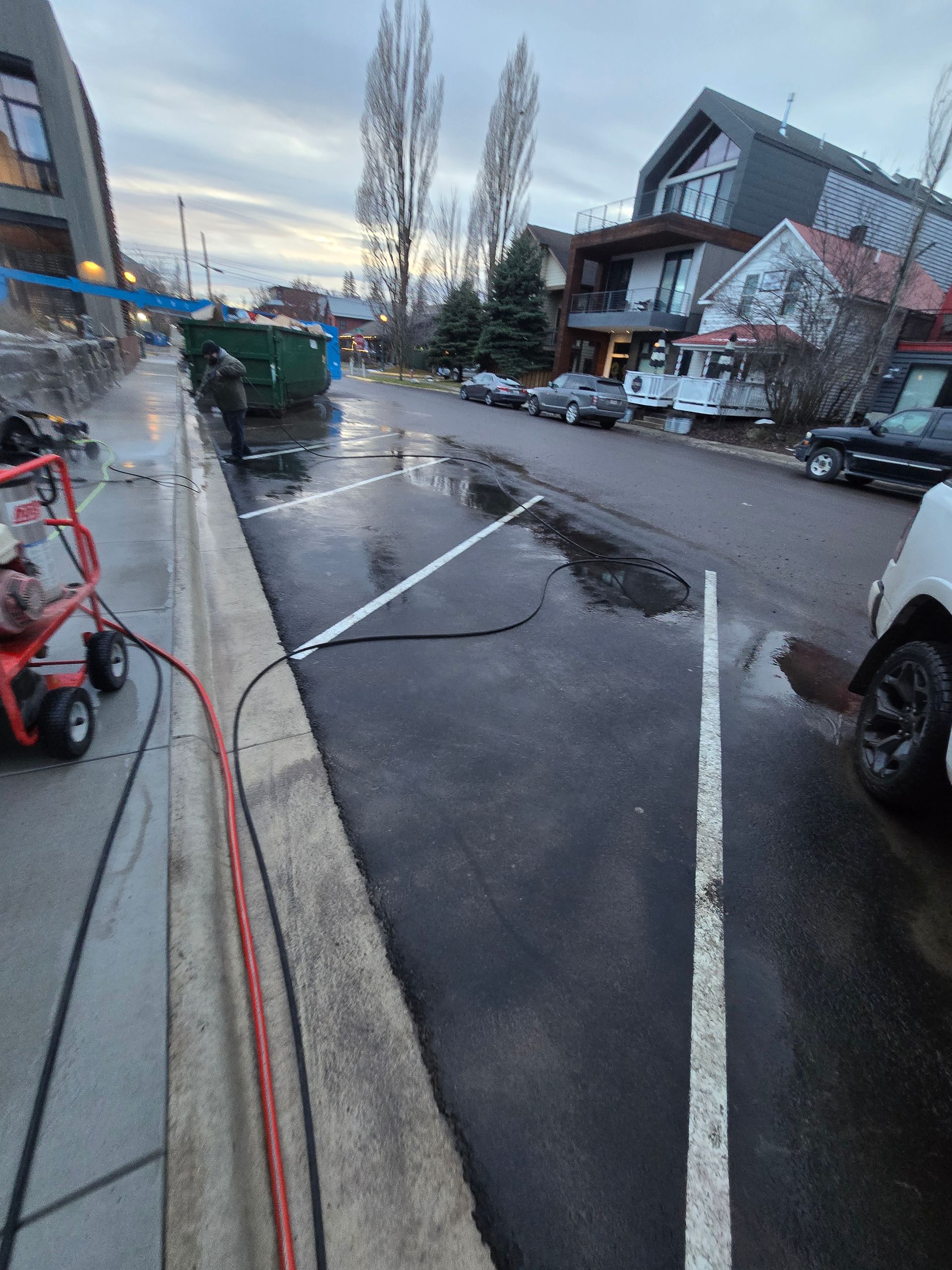 A street scene showing a wet parking lane, white parking lines, a green dumpster, and construction equipment on the curb.