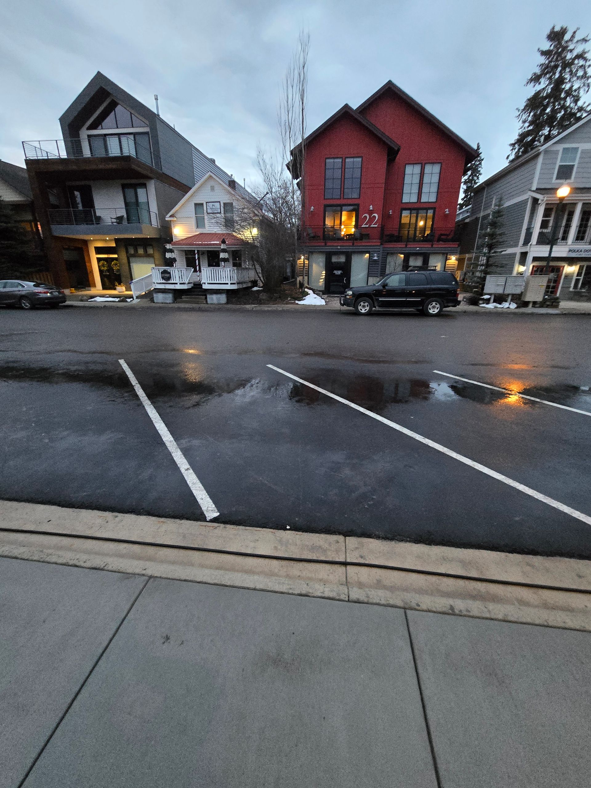 Street view of buildings, including a prominent red house, behind diagonal parking spaces on a paved road at dusk.