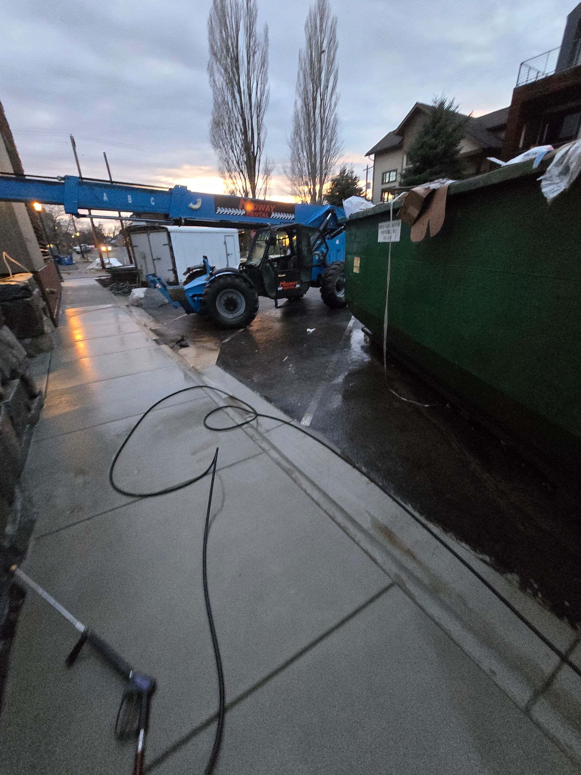 A blue telehandler with an extended boom parked next to a large green dumpster on a concrete street at dusk.