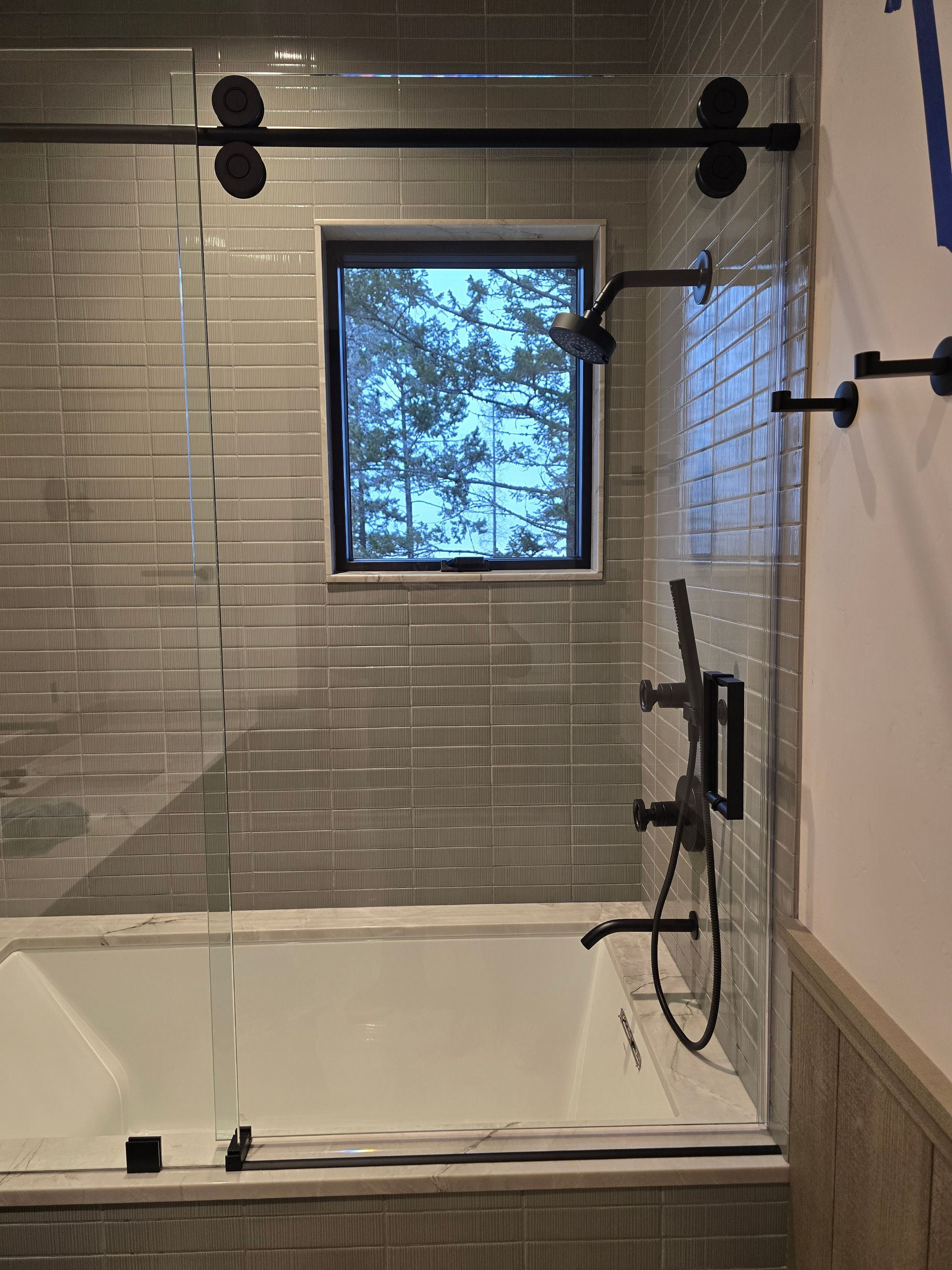 A modern bathtub with a window, gray tile wall, and glass shower door with matte black hardware and a handheld shower head.