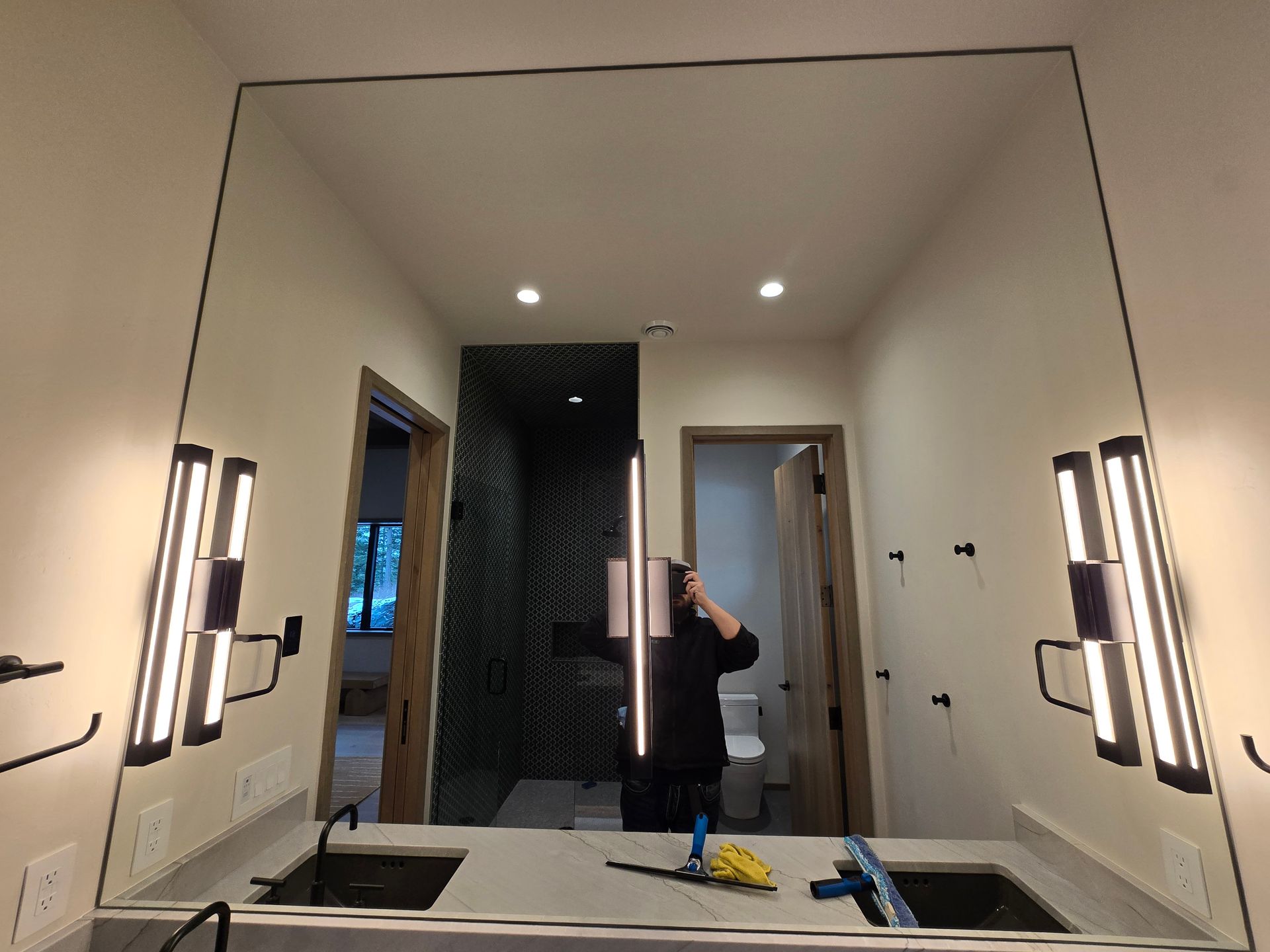 A bathroom vanity with a large mirror, two modern vertical wall lights, double sinks, and cleaning supplies on the counter.