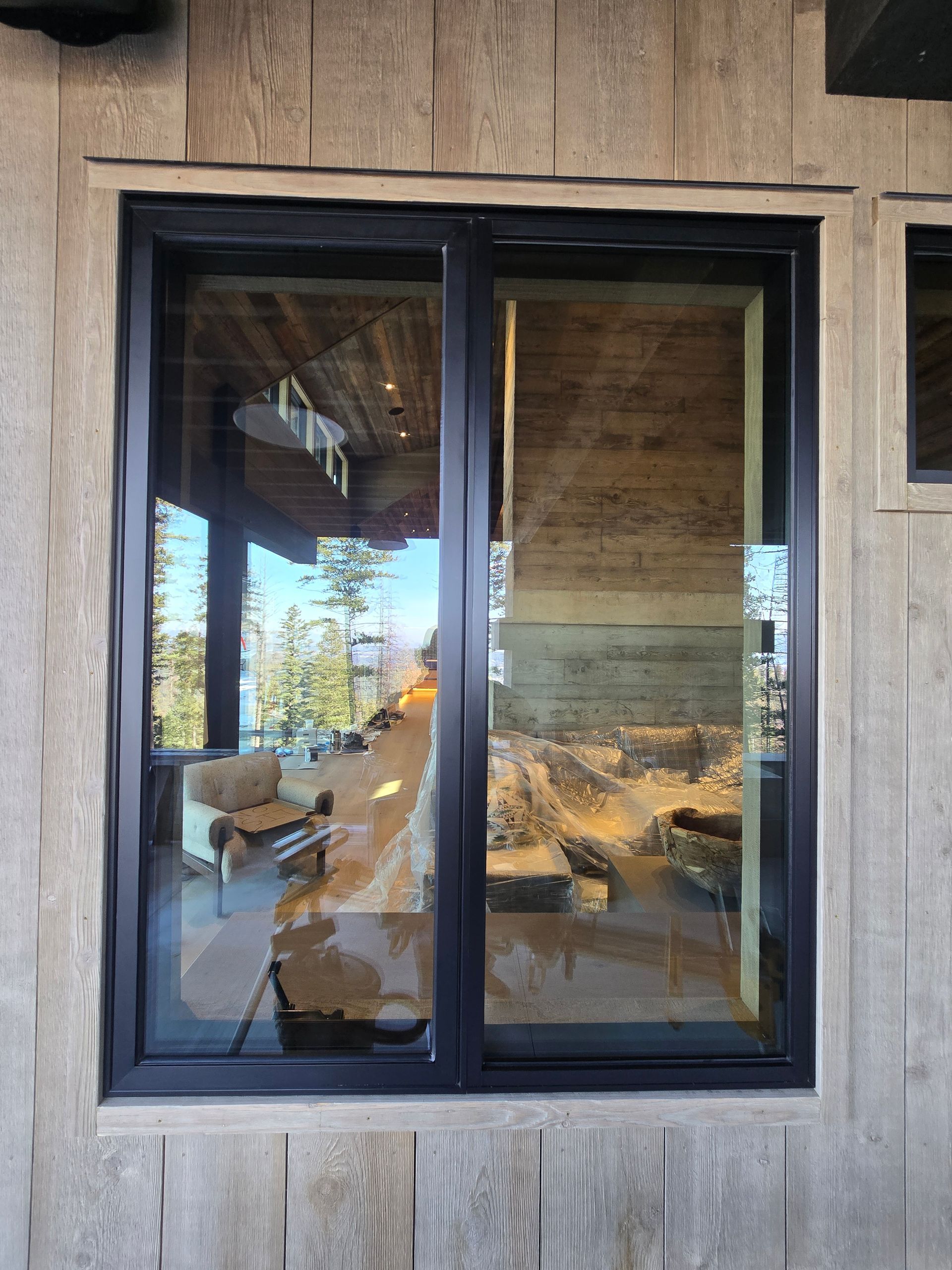 A black-framed window on a rustic, light wood-paneled wall, reflecting the interior and outdoor landscape.