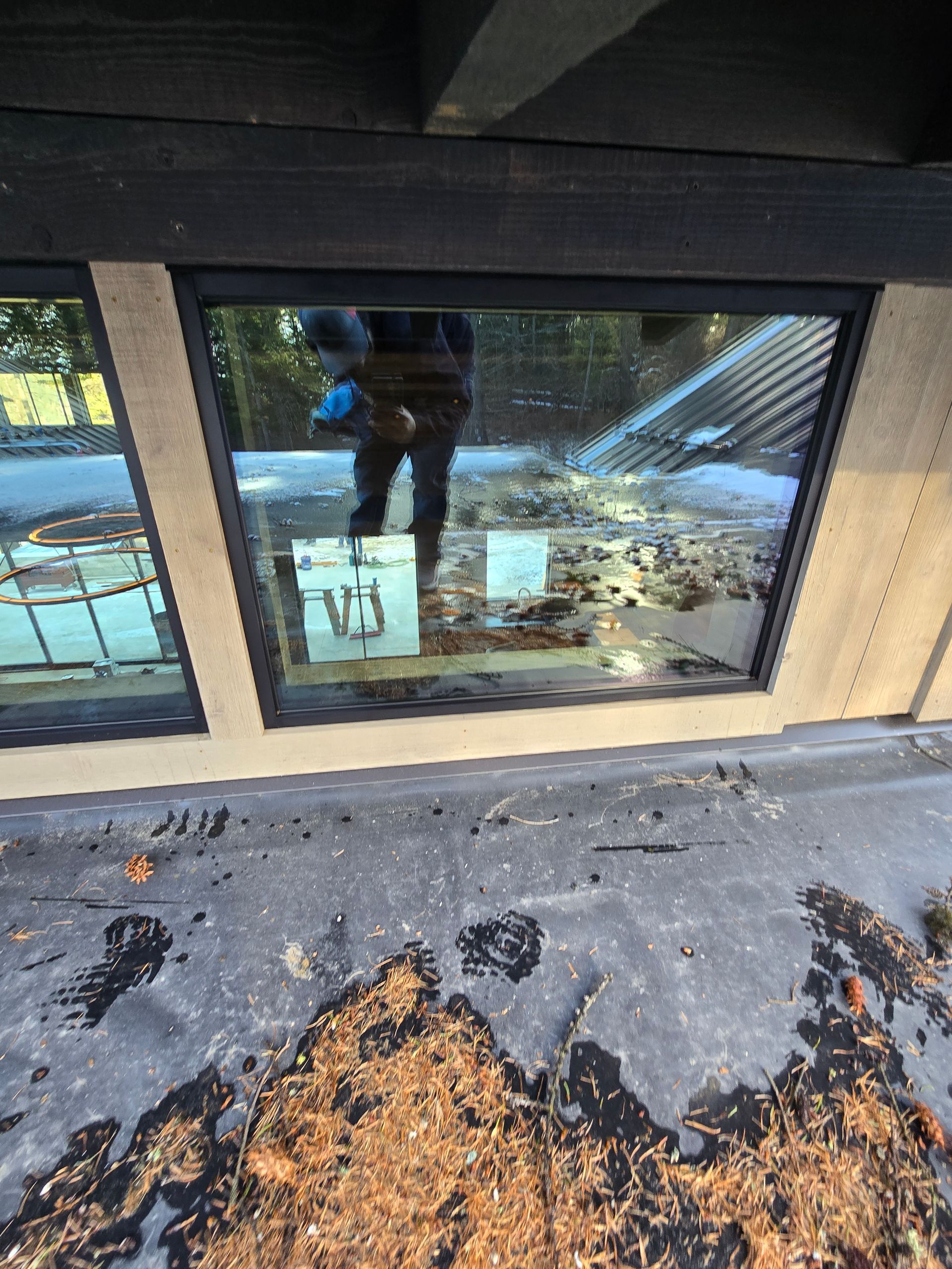 A view of a roof surface covered in debris, seen through a glass window showing the reflection of a person taking a photo.