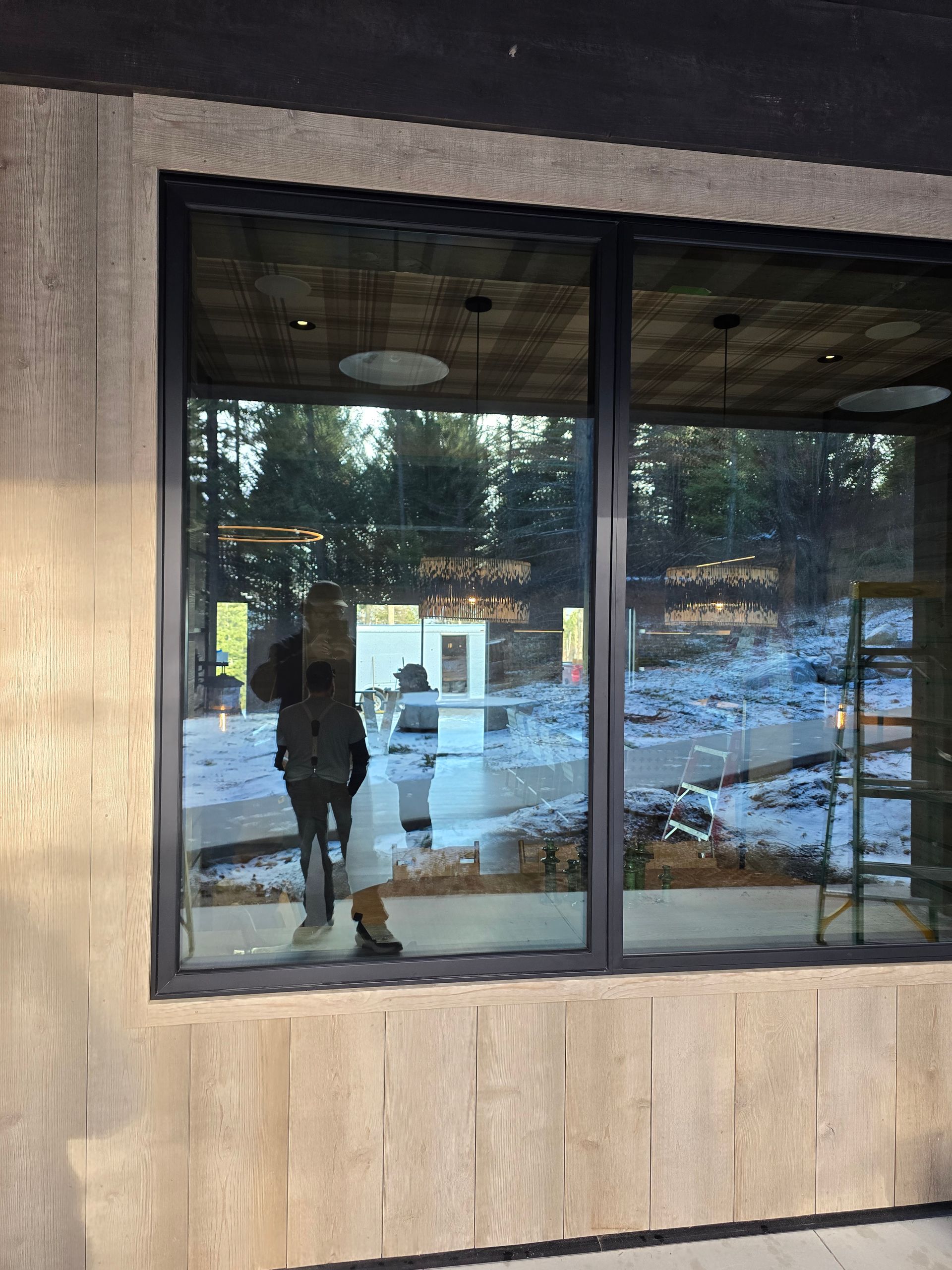 Reflective glass windows of a building under construction, showing a person taking a photo and snow on the ground outside.