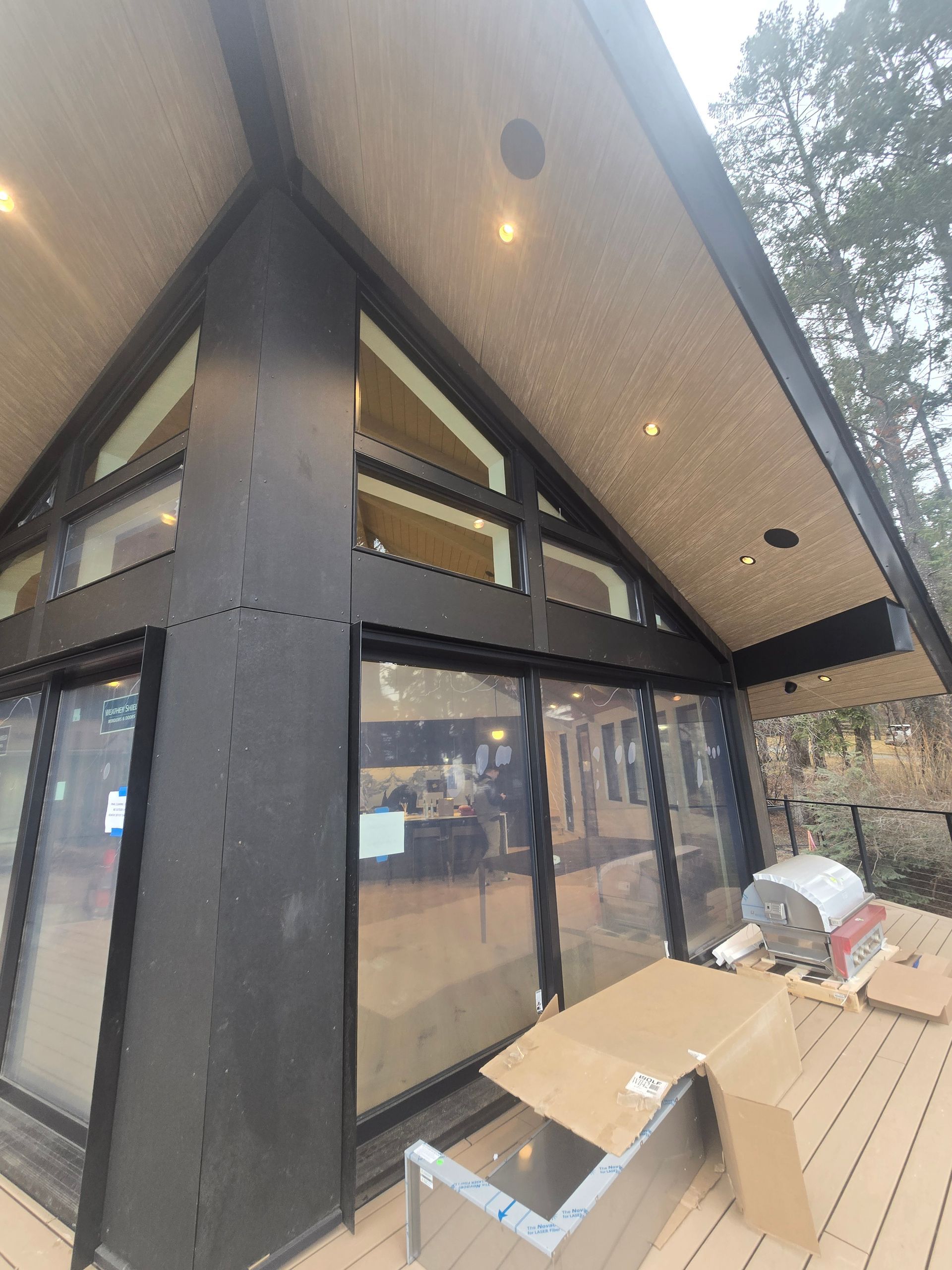 A wooden deck with a grill and boxes sits outside a modern cabin with dark metal framing and large glass windows.