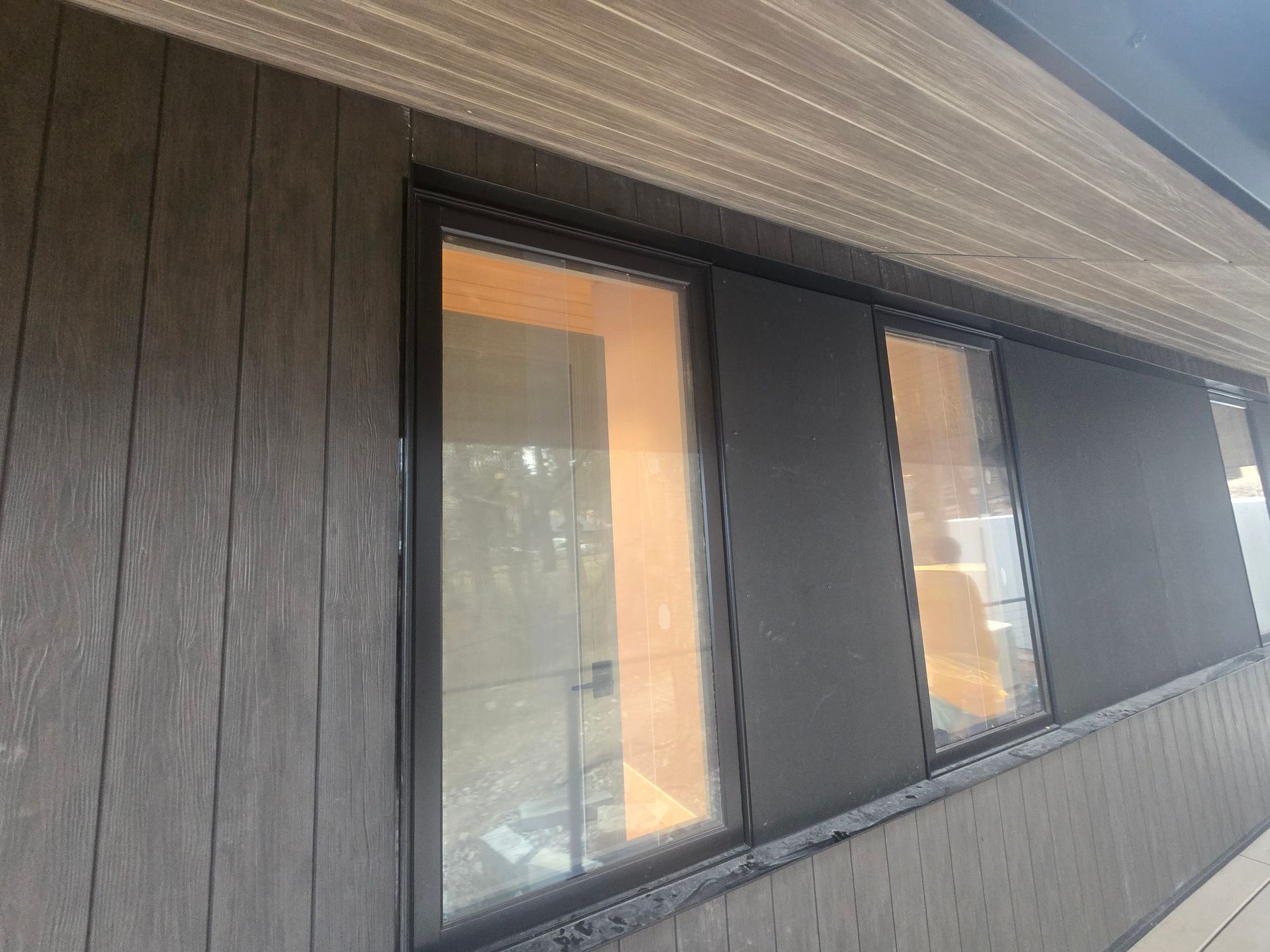 Exterior view of a dark wood-paneled wall with two large windows and a beige soffit overhead.