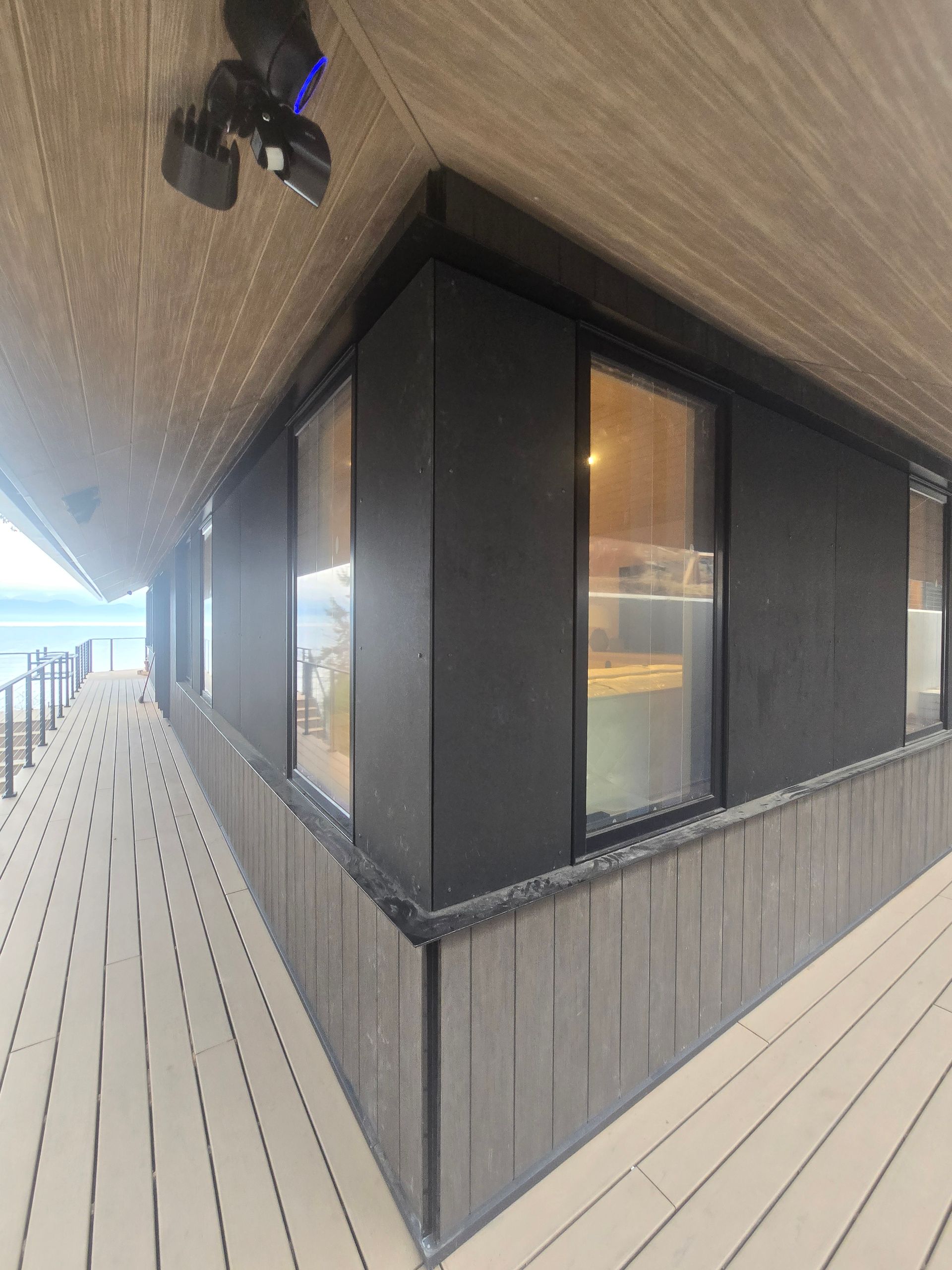 A low-angle view of a building corner with black exterior walls, dark windows, and a wood-slat deck overlooking a lake.