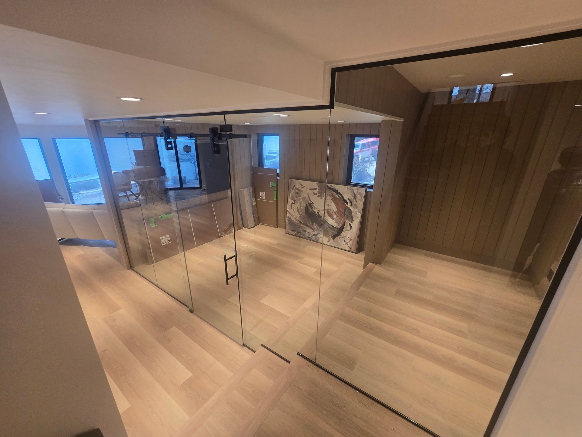Modern basement interior with light wood flooring, a glass-enclosed room, and wood-paneled walls under recessed lighting.