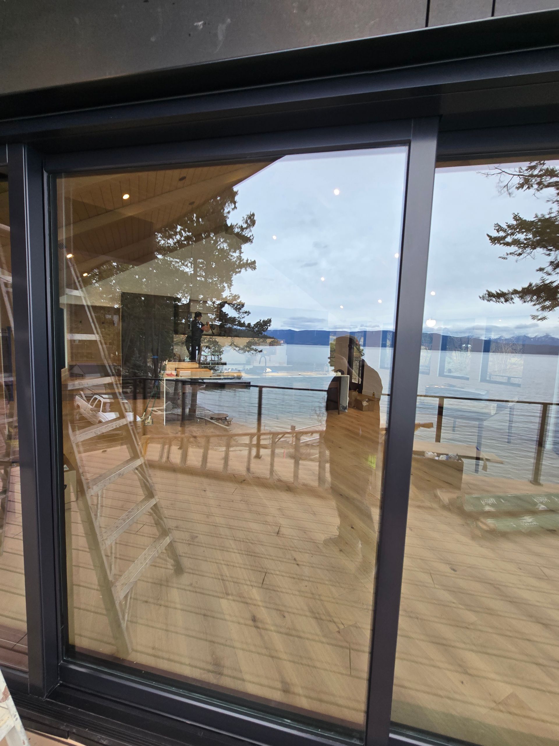 A view through a large glass sliding door showing a wooden deck, a lake, and trees, with a person reflected in the glass.