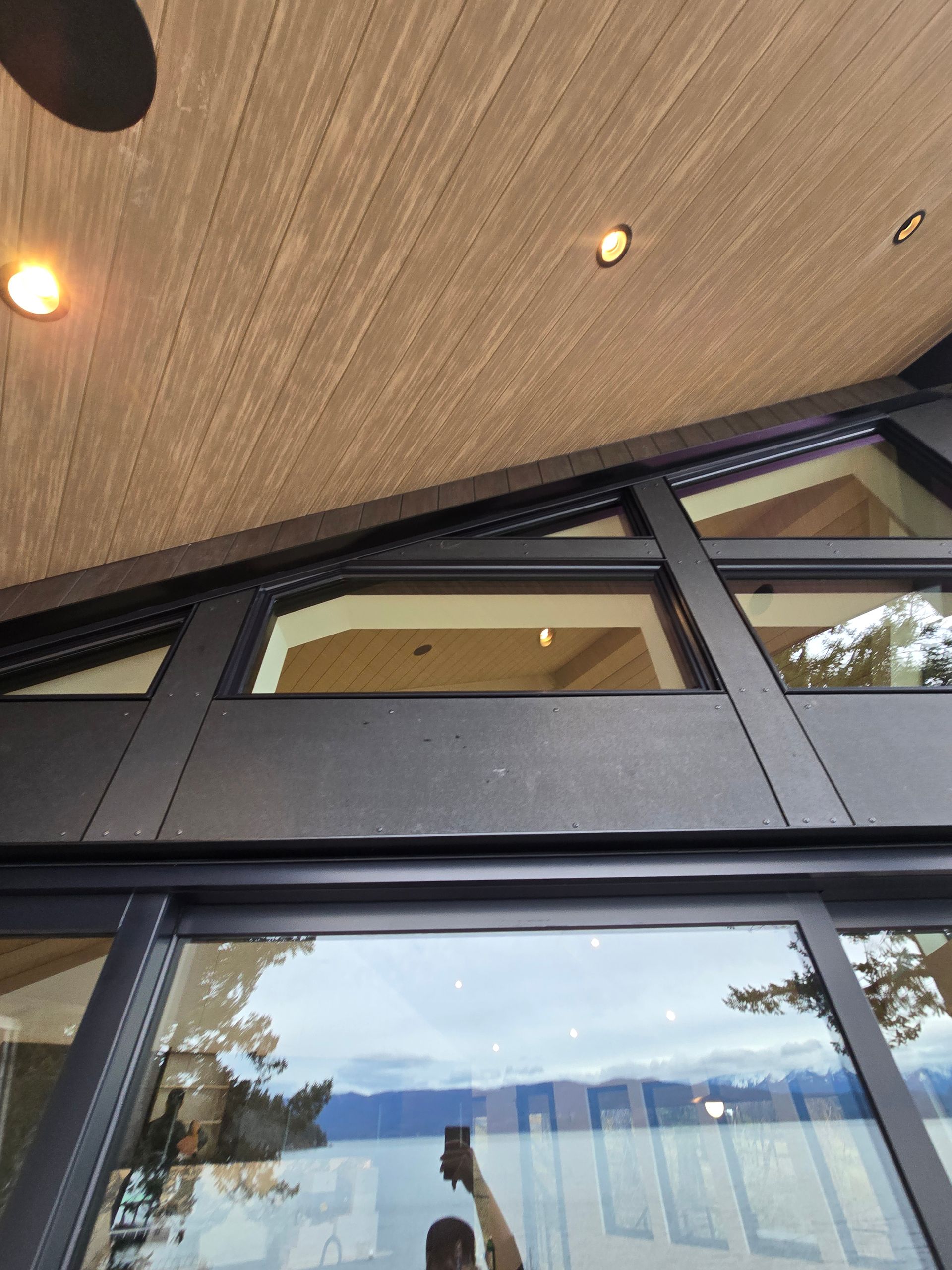 A low-angle view of a dark metal window frame and wooden ceiling with recessed lighting overlooking a lake.