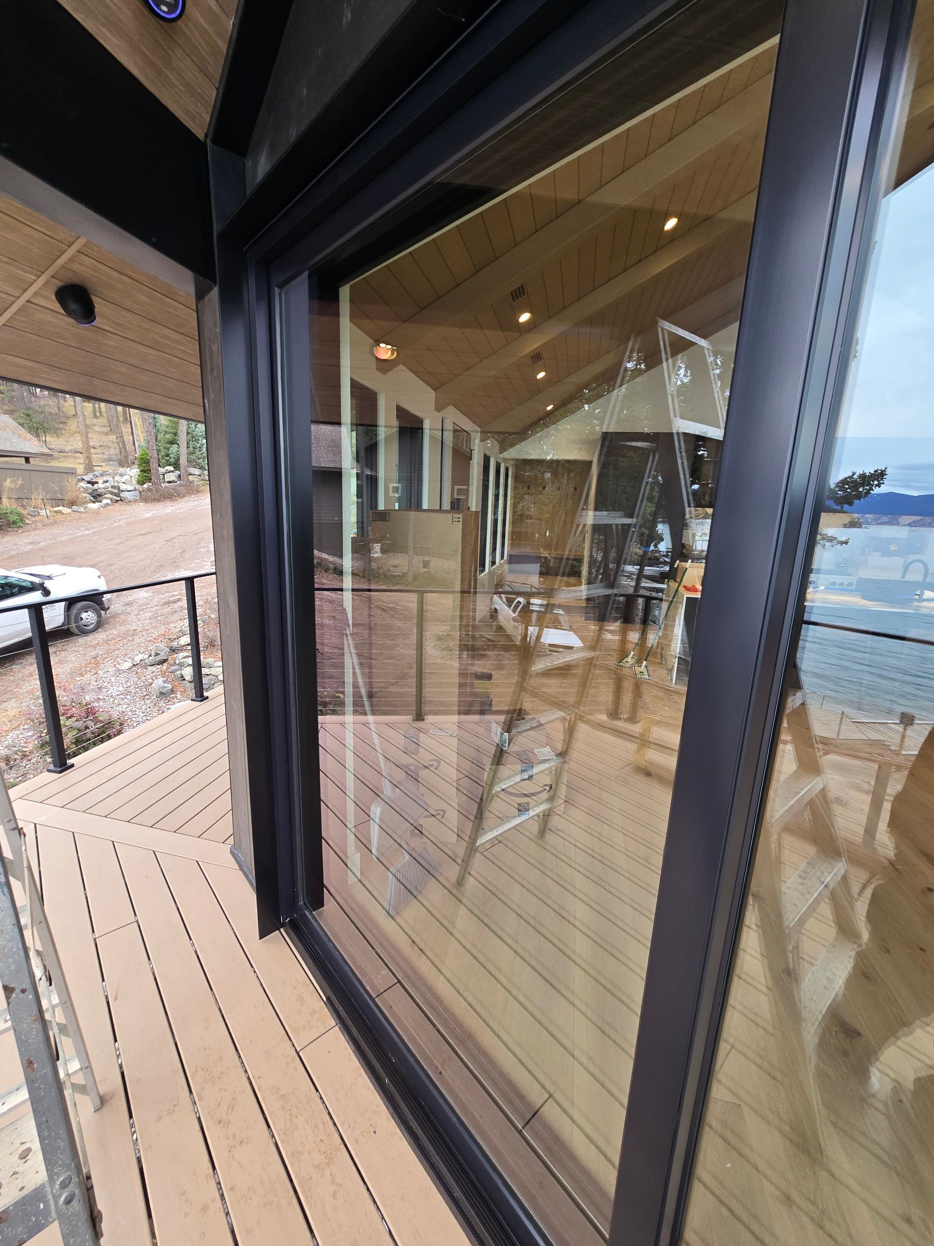 Large black-framed glass sliding doors open from a wooden deck onto a modern, unfinished indoor space overlooking a lake.
