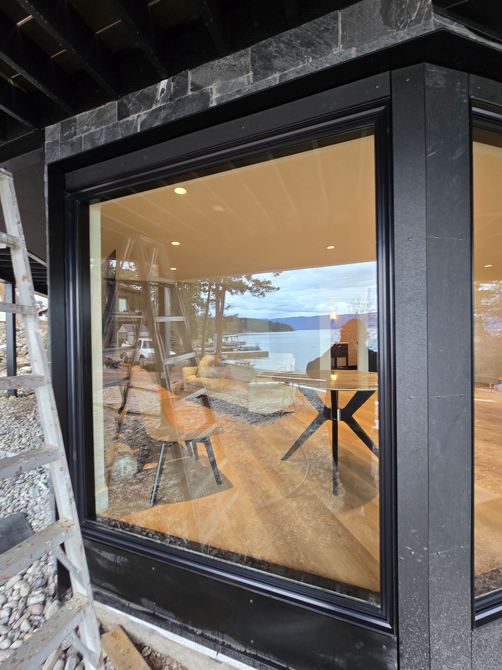 A large, black-framed bay window showing a reflection of a modern room with furniture and a scenic view of the water.
