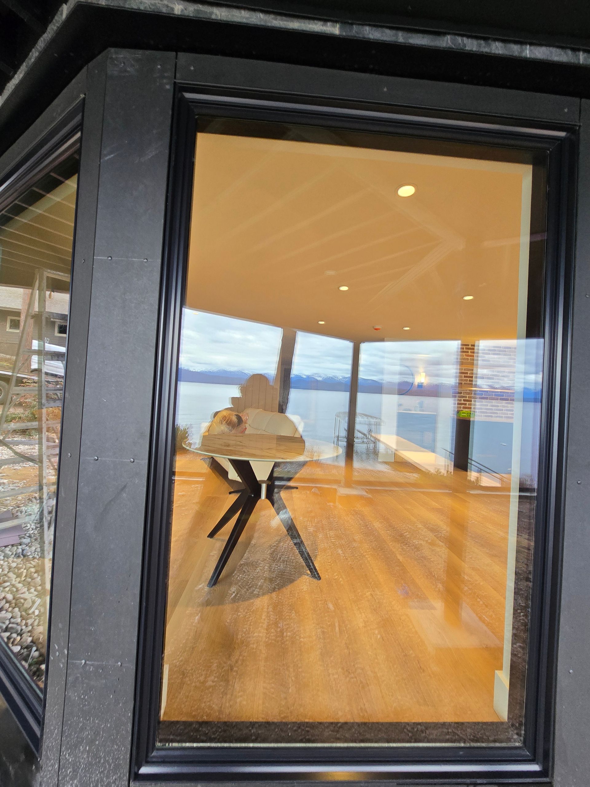 A window view of a minimalist wooden table in a bright, modern room overlooking a large body of water and distant mountains.