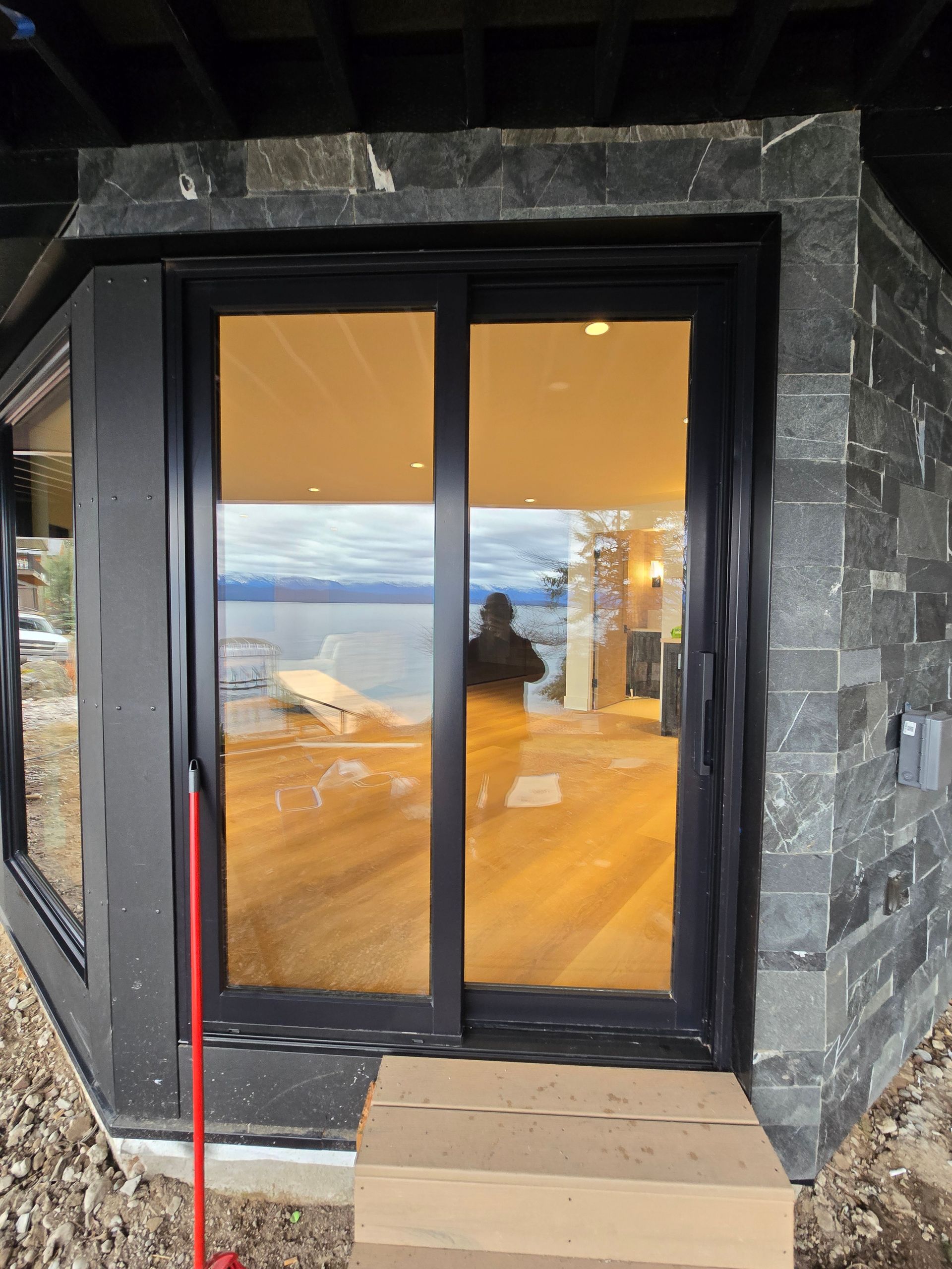Black sliding glass door set into dark stone siding, with a view of a lake and interior reflections through the glass.