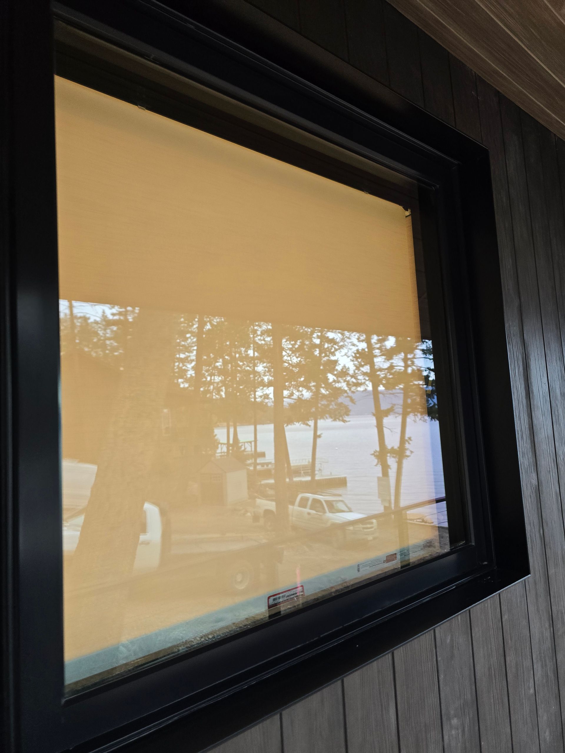 A close-up view of a dark-framed window reflecting a lakeside scene with trees and vehicles.