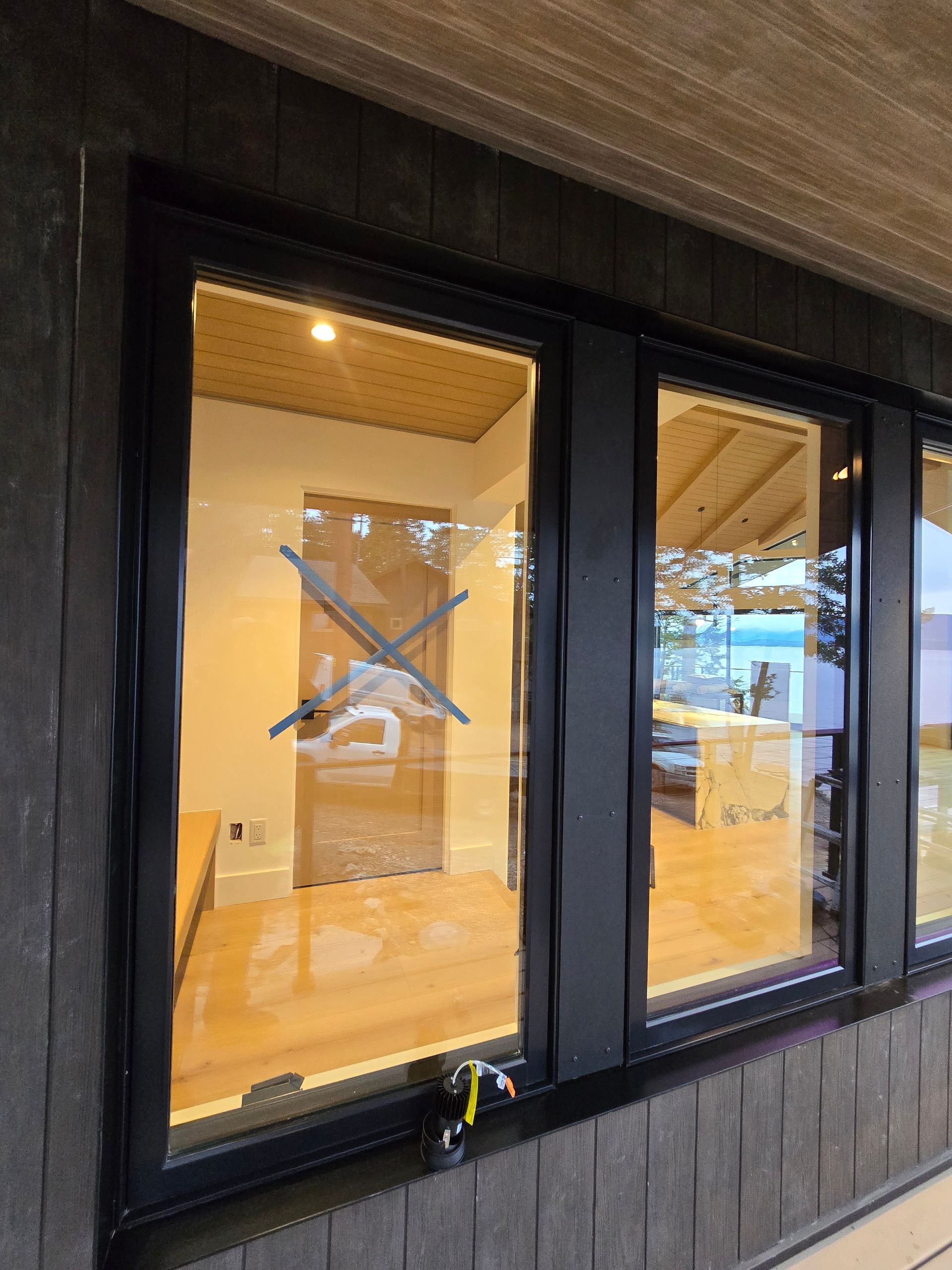 Black-framed windows with an X-shaped tape marking on a dark shingled exterior wall, reflecting an indoor space.