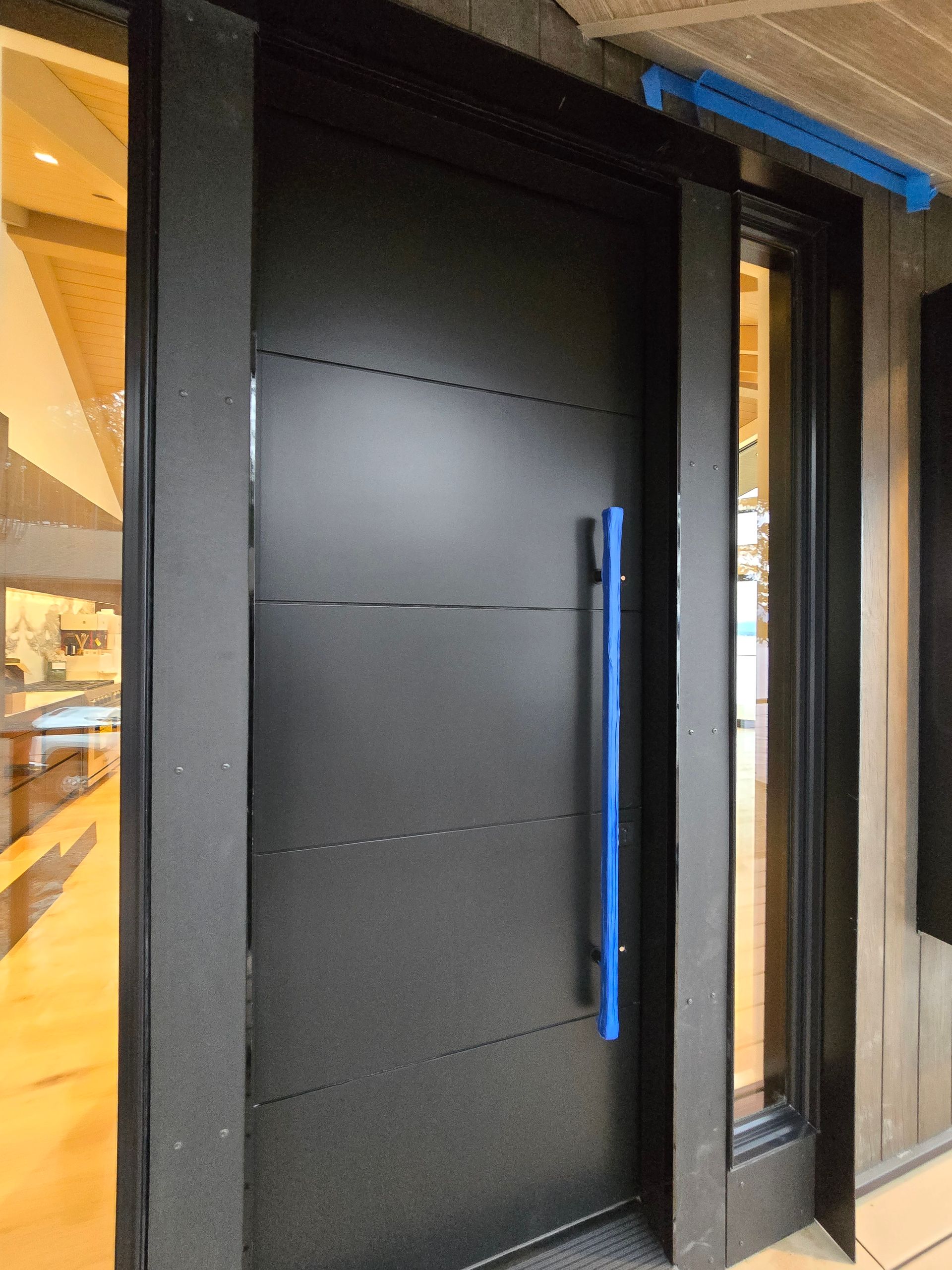 A black, modern front door with horizontal lines and a prominent blue vertical handle next to a narrow window panel.