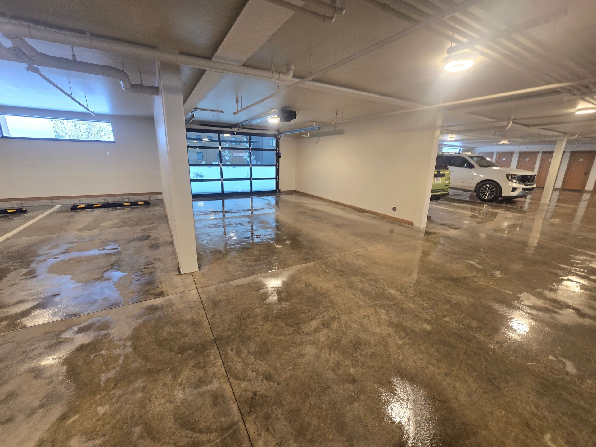 A wide, brightly lit underground parking garage with wet concrete floors, white walls, and a white SUV parked in back.
