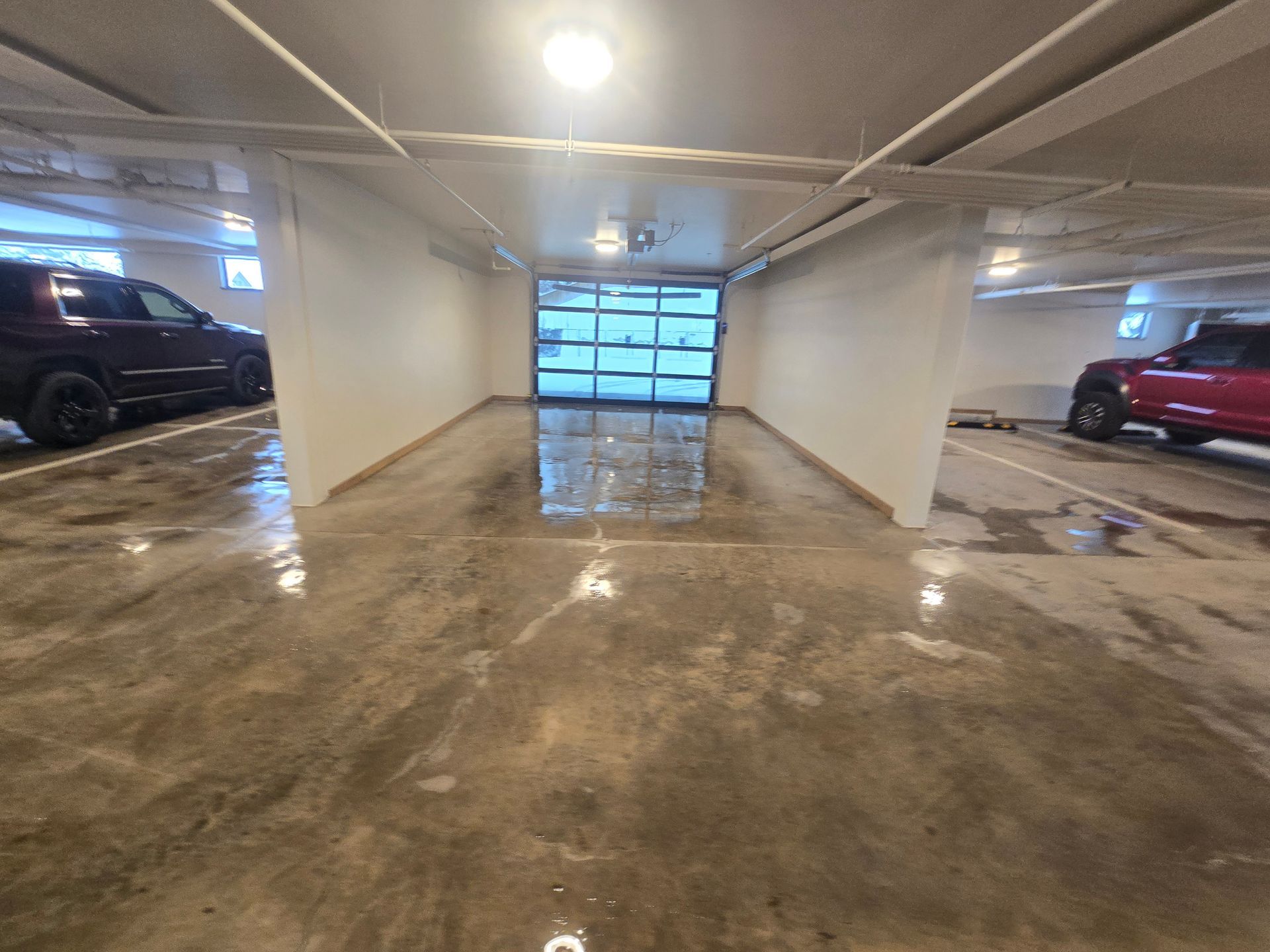 A dimly lit indoor parking garage with a wet concrete floor, showing parked vehicles on either side of a central lane.