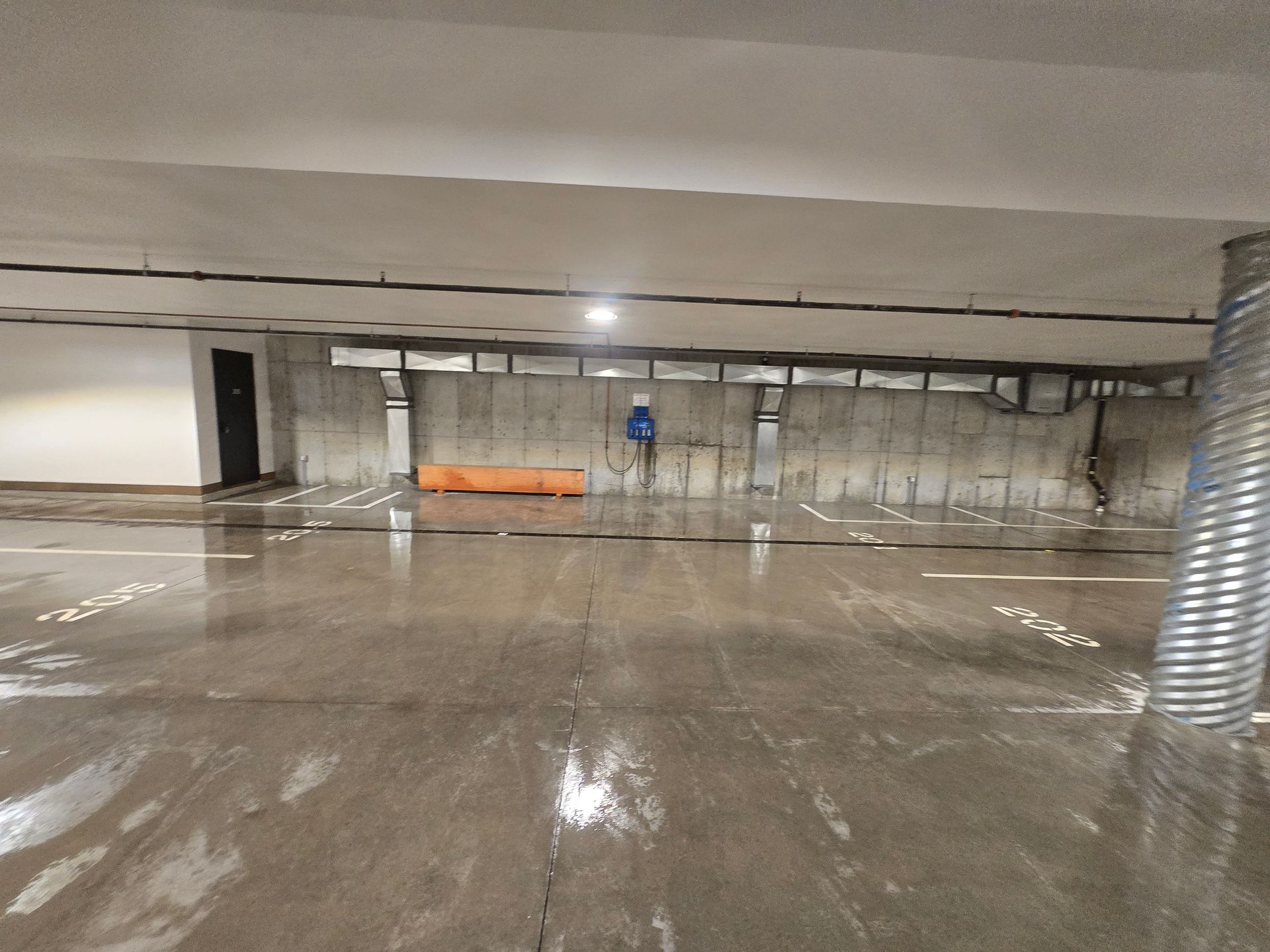 A wide view of a dimly lit, empty underground parking garage with a wet concrete floor, exposed ductwork, and white walls.