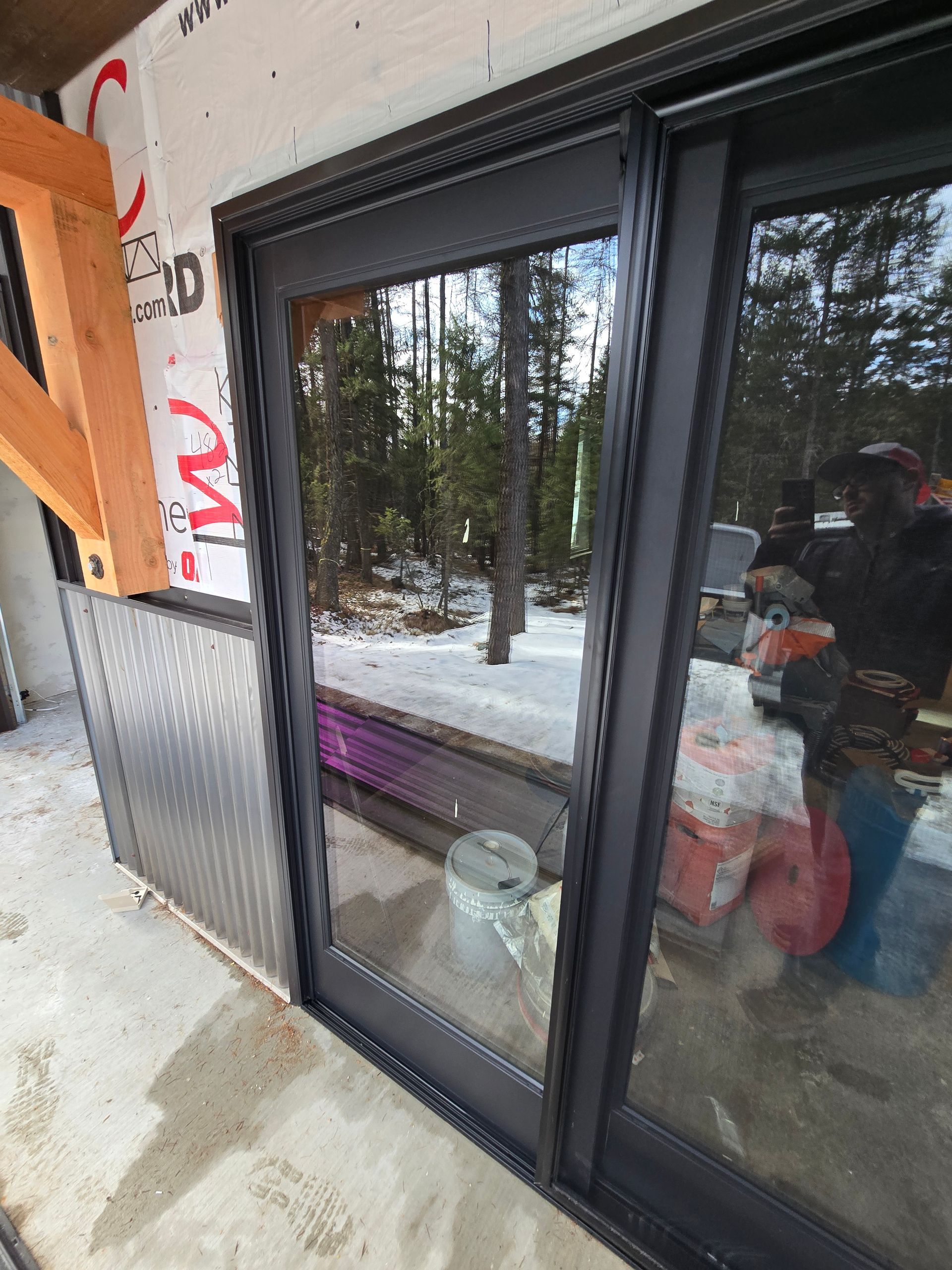 A dark-framed sliding glass door installed in an unfinished exterior wall with wood framing and corrugated metal siding.
