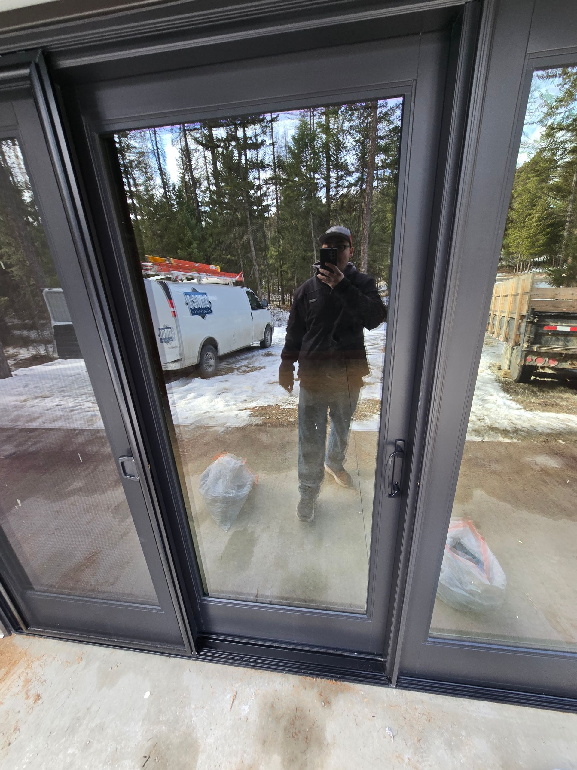 A person taking a selfie in the reflection of a set of dark-framed glass doors outdoors on a snowy day.
