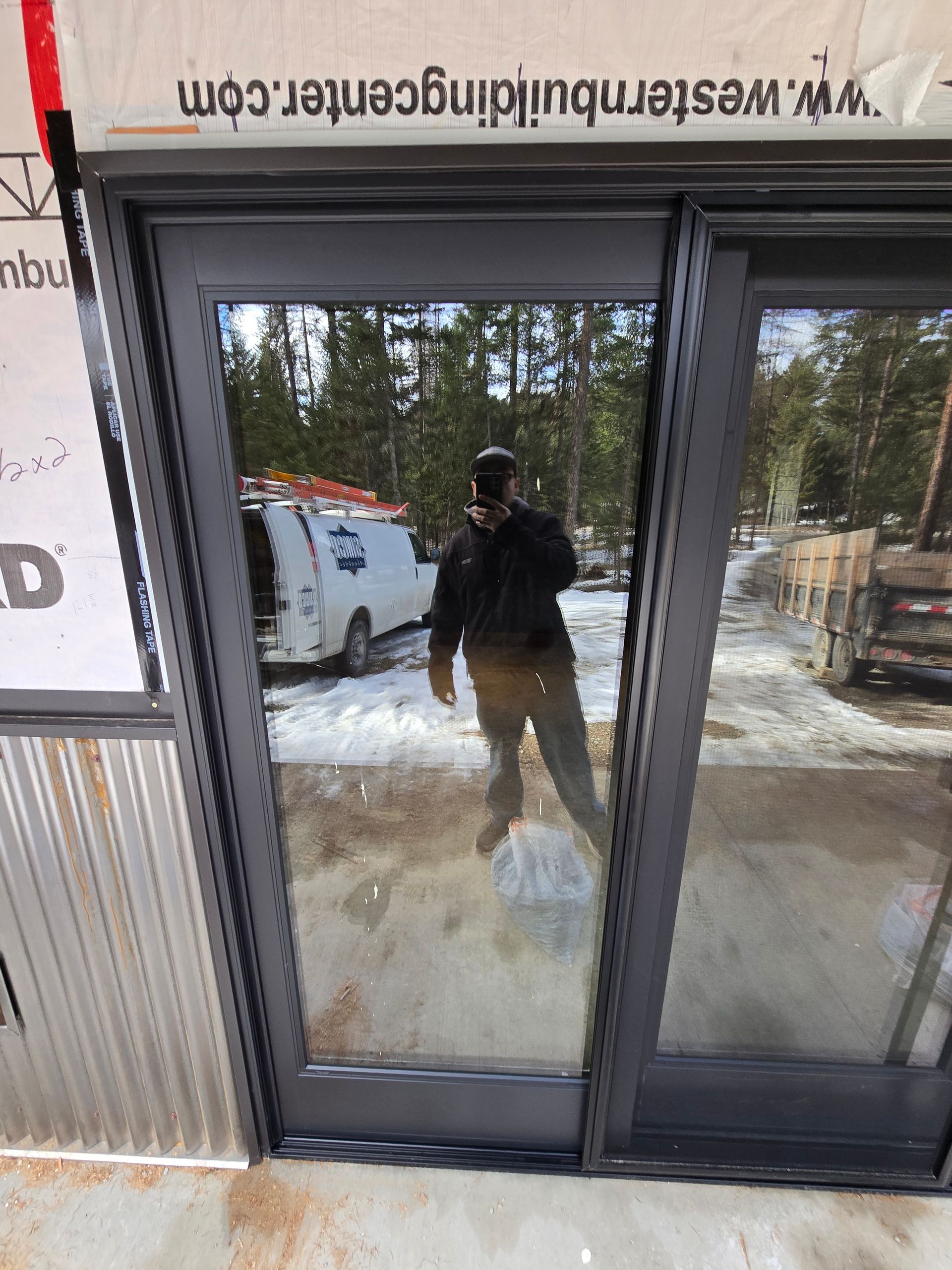 A person standing in front of a newly installed dark-framed sliding glass door at a snowy construction site.