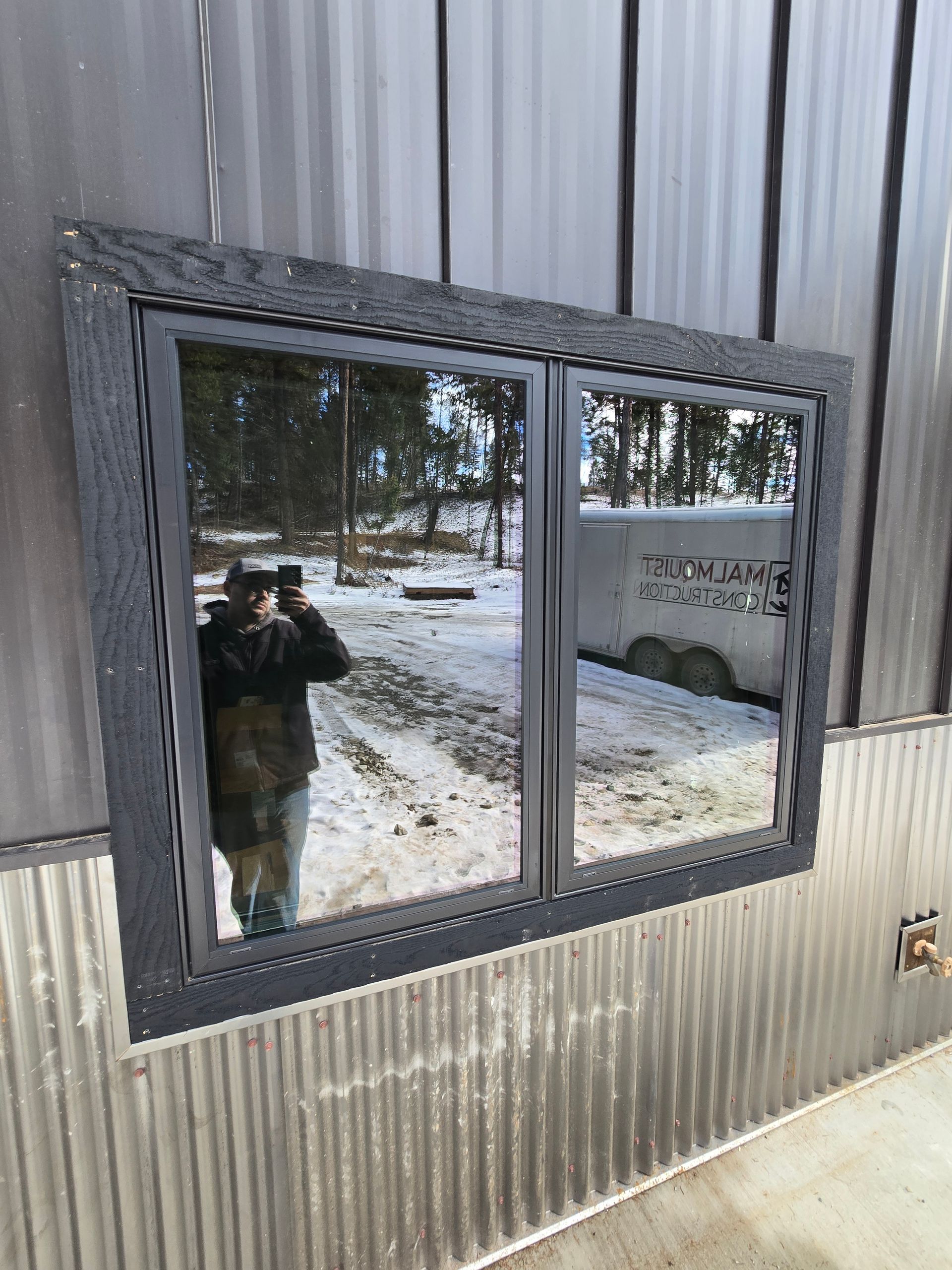 A person taking a photo in the reflection of a dark-framed window on a building with corrugated metal siding.