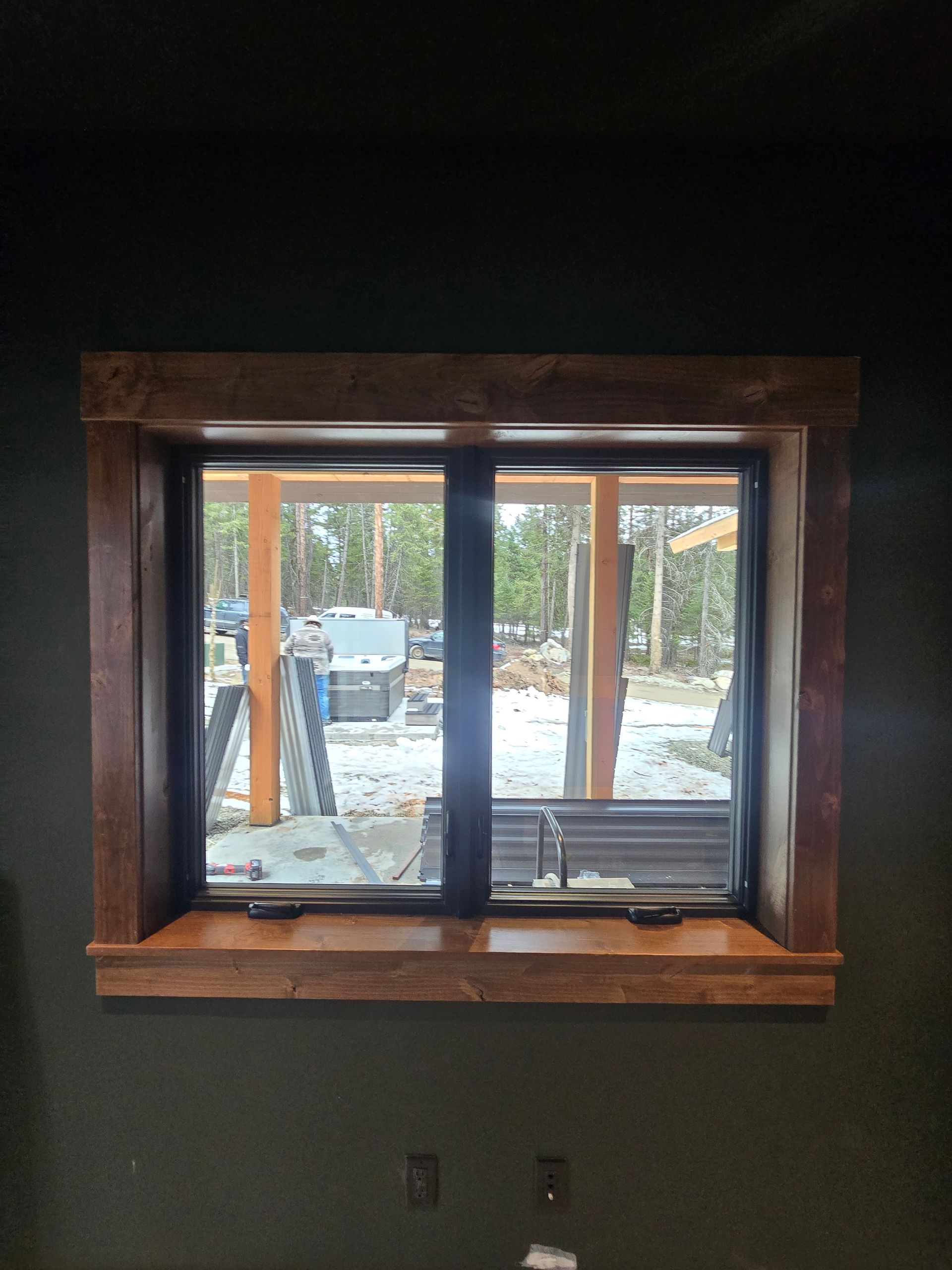 A dark-walled room features a window with a natural wood frame and sill, overlooking a snow-covered, wooded exterior.