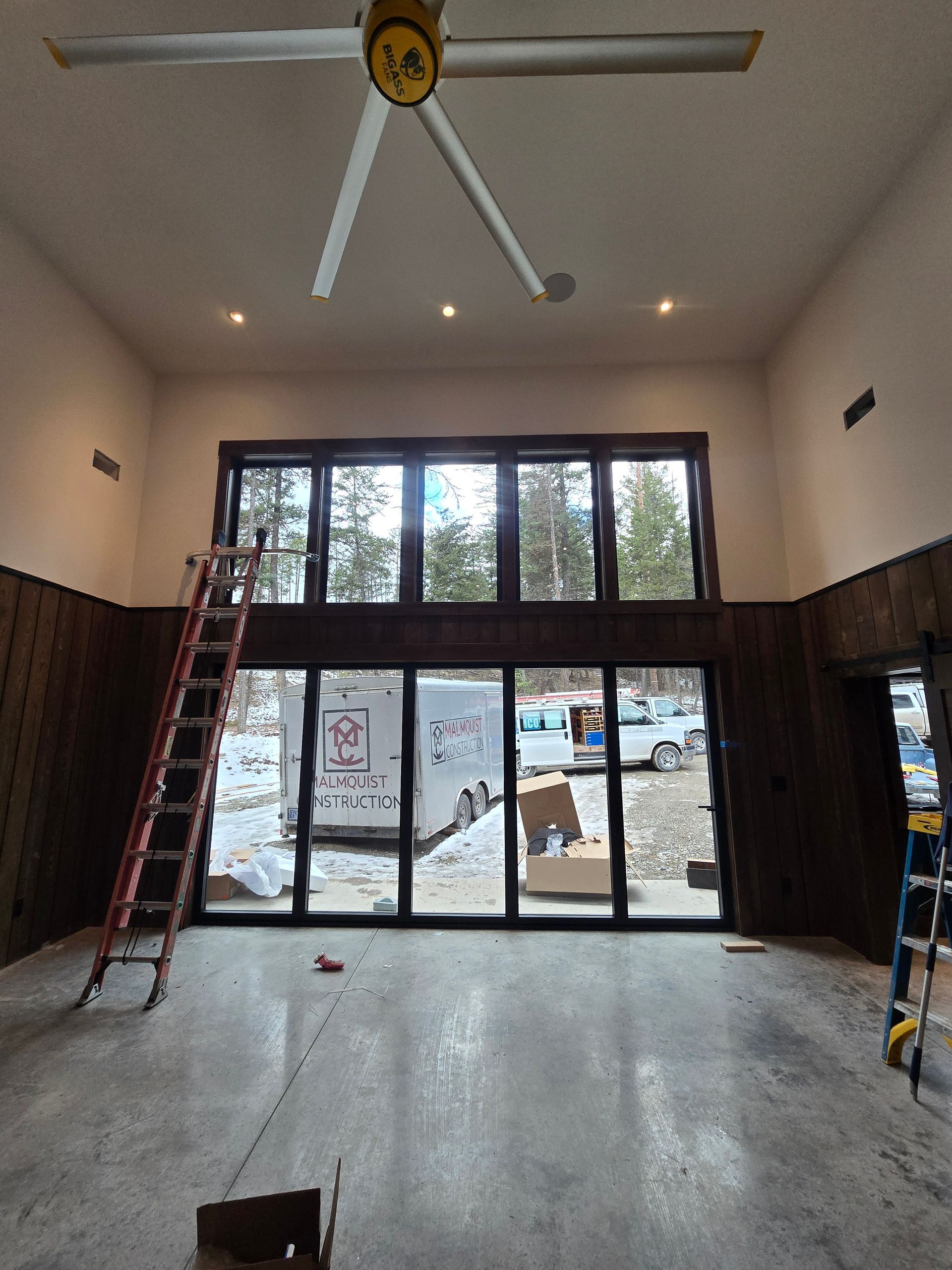 Interior of a room under construction with a large, two-story grid window wall, ladders, and concrete flooring.