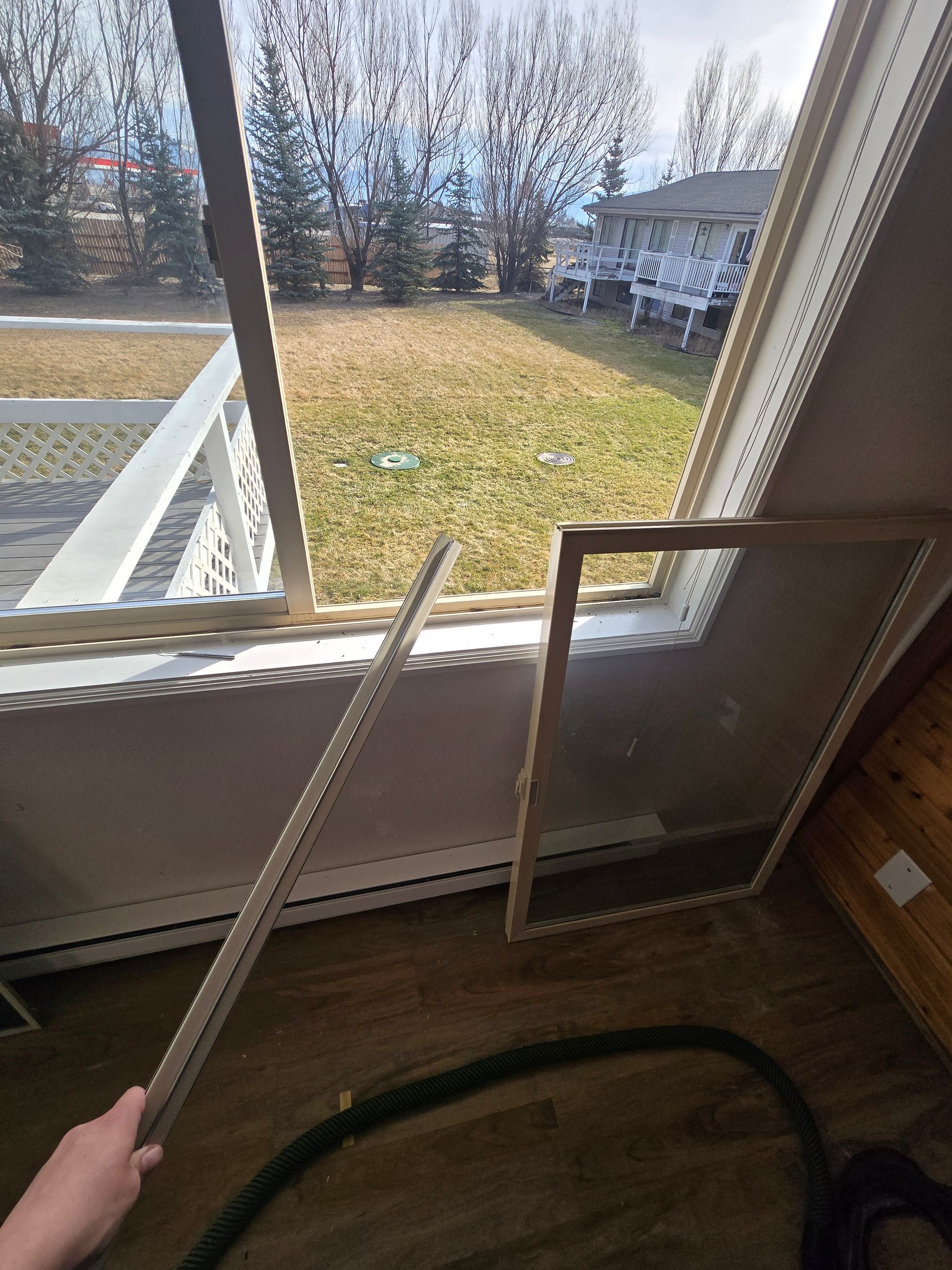 A person holding a detached window screen strip next to an open window frame, with a backyard visible through the glass.