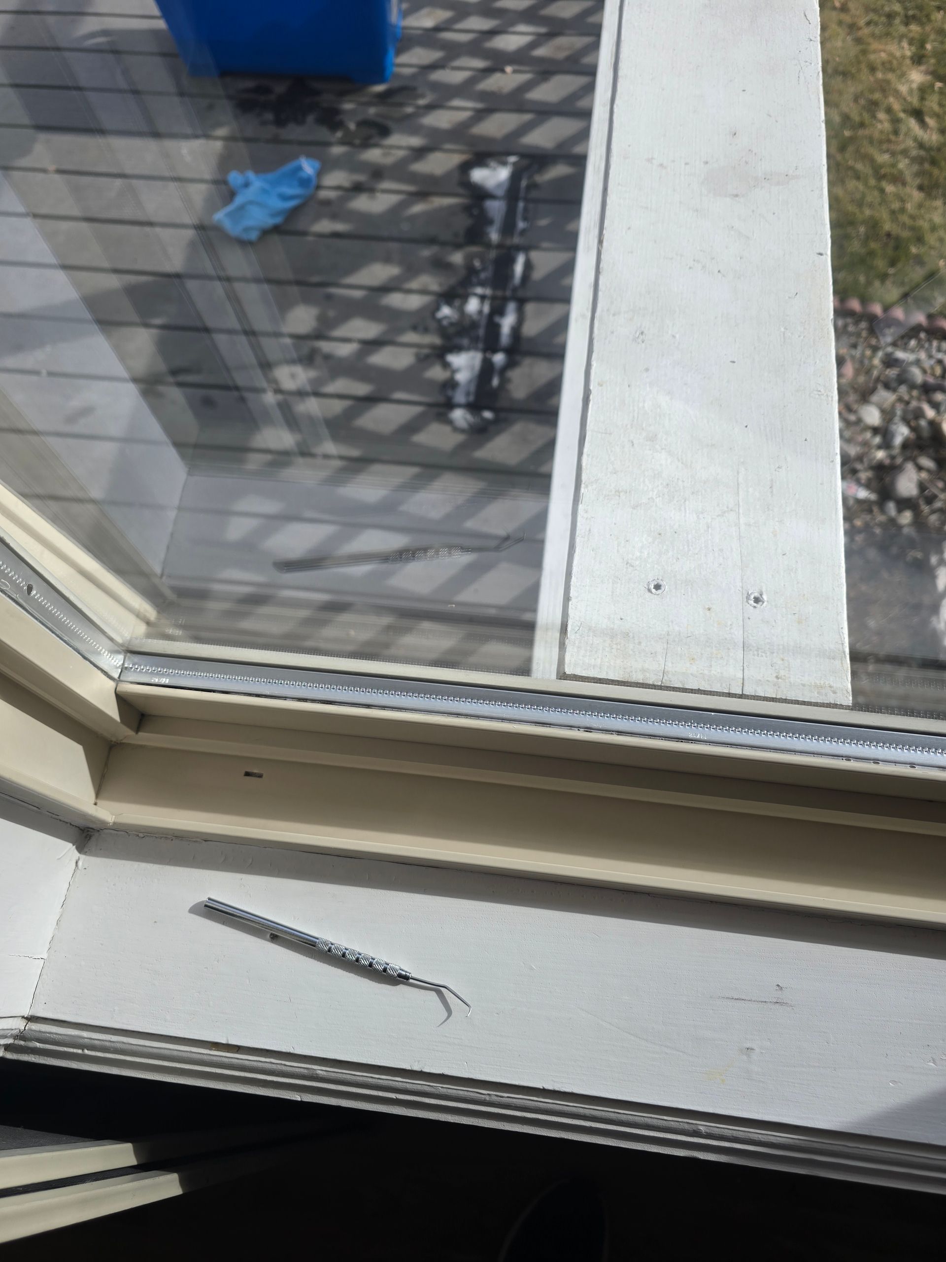 A silver spring-loaded window balance mechanism sits on a window sill with a backyard deck visible through the glass.
