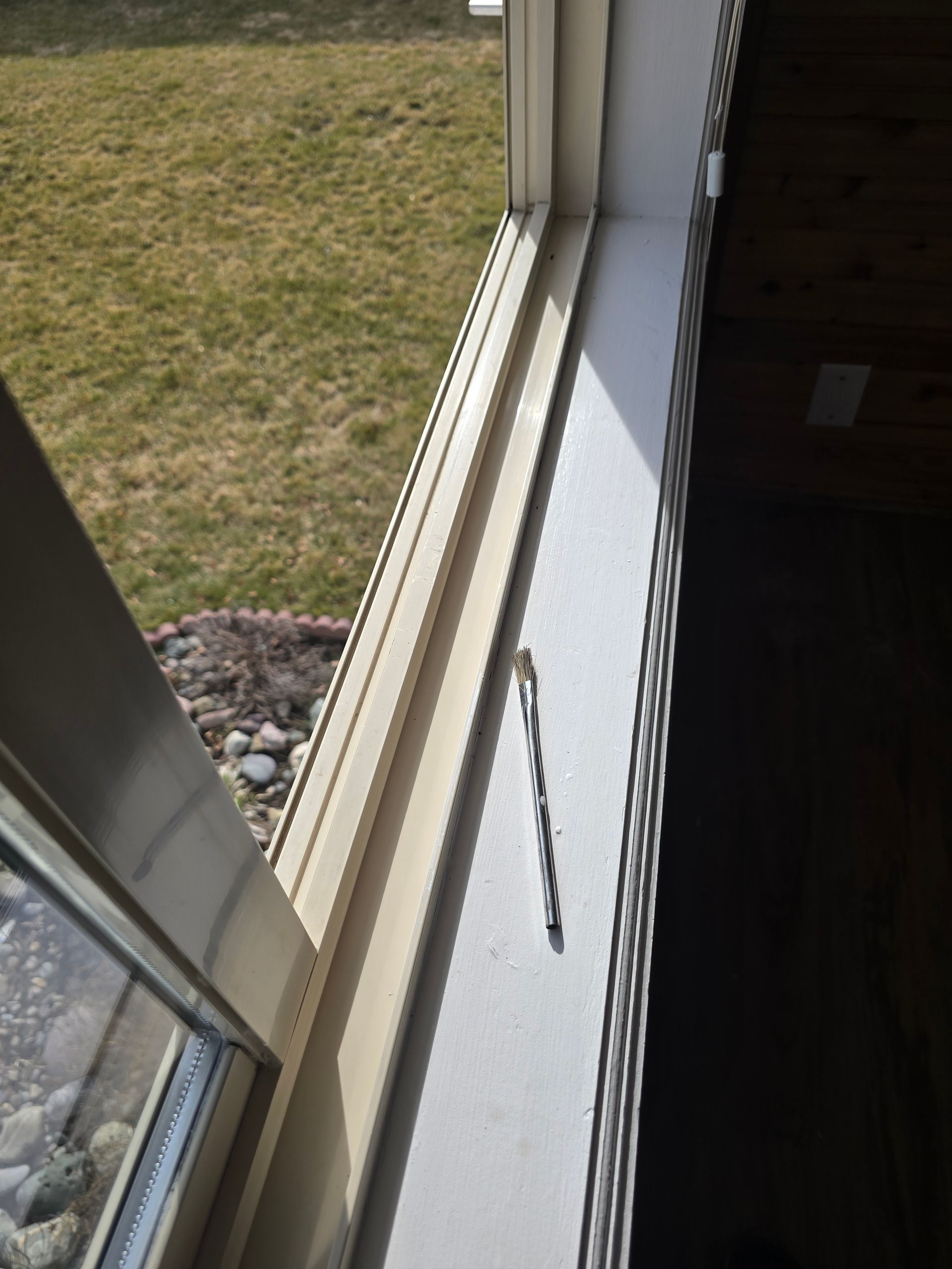 A drill bit rests on a white window sill next to a window overlooking a yard.