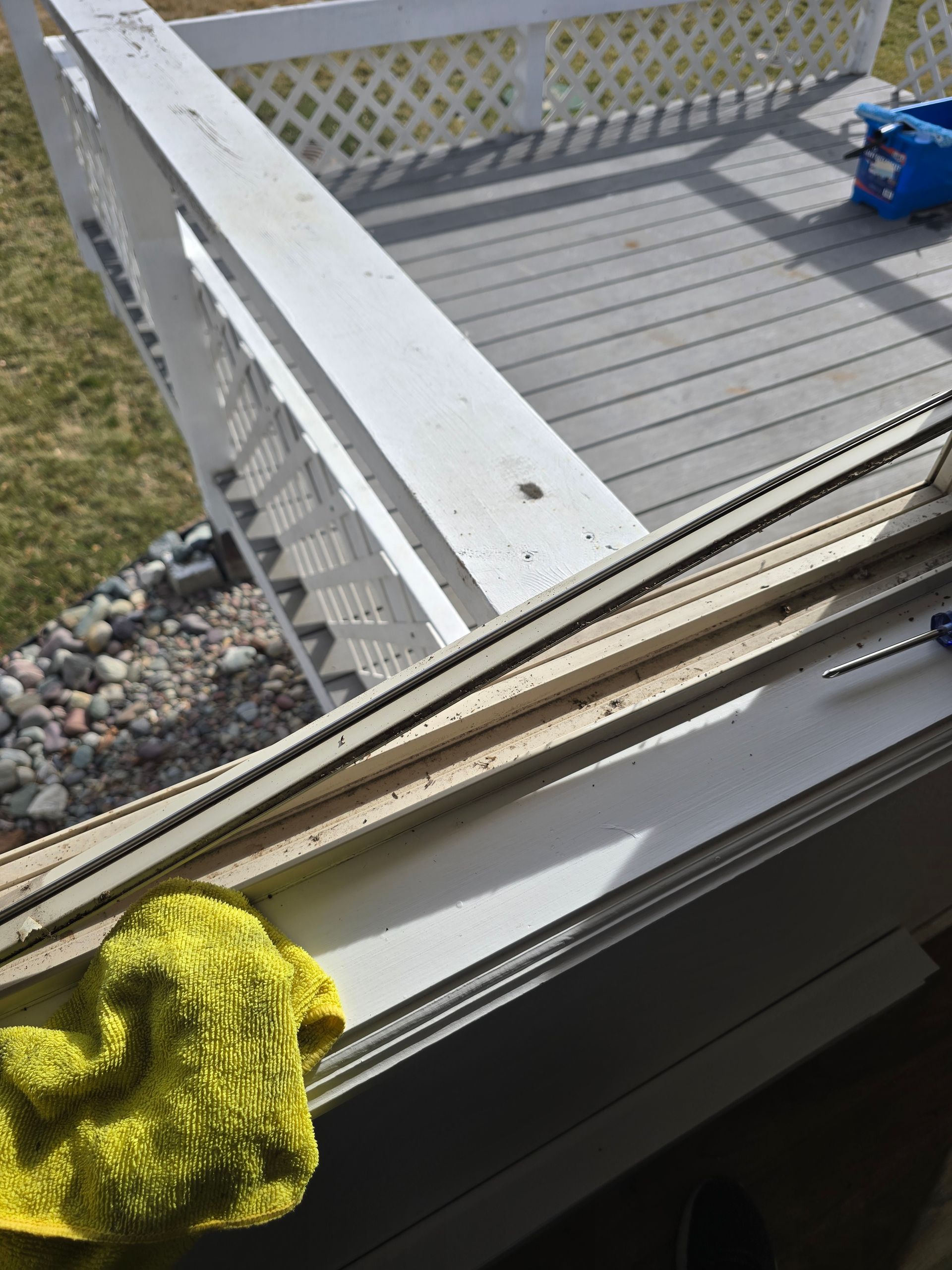 A yellow cloth rests on a white windowsill next to a removed screen frame, overlooking a grey deck with a railing.