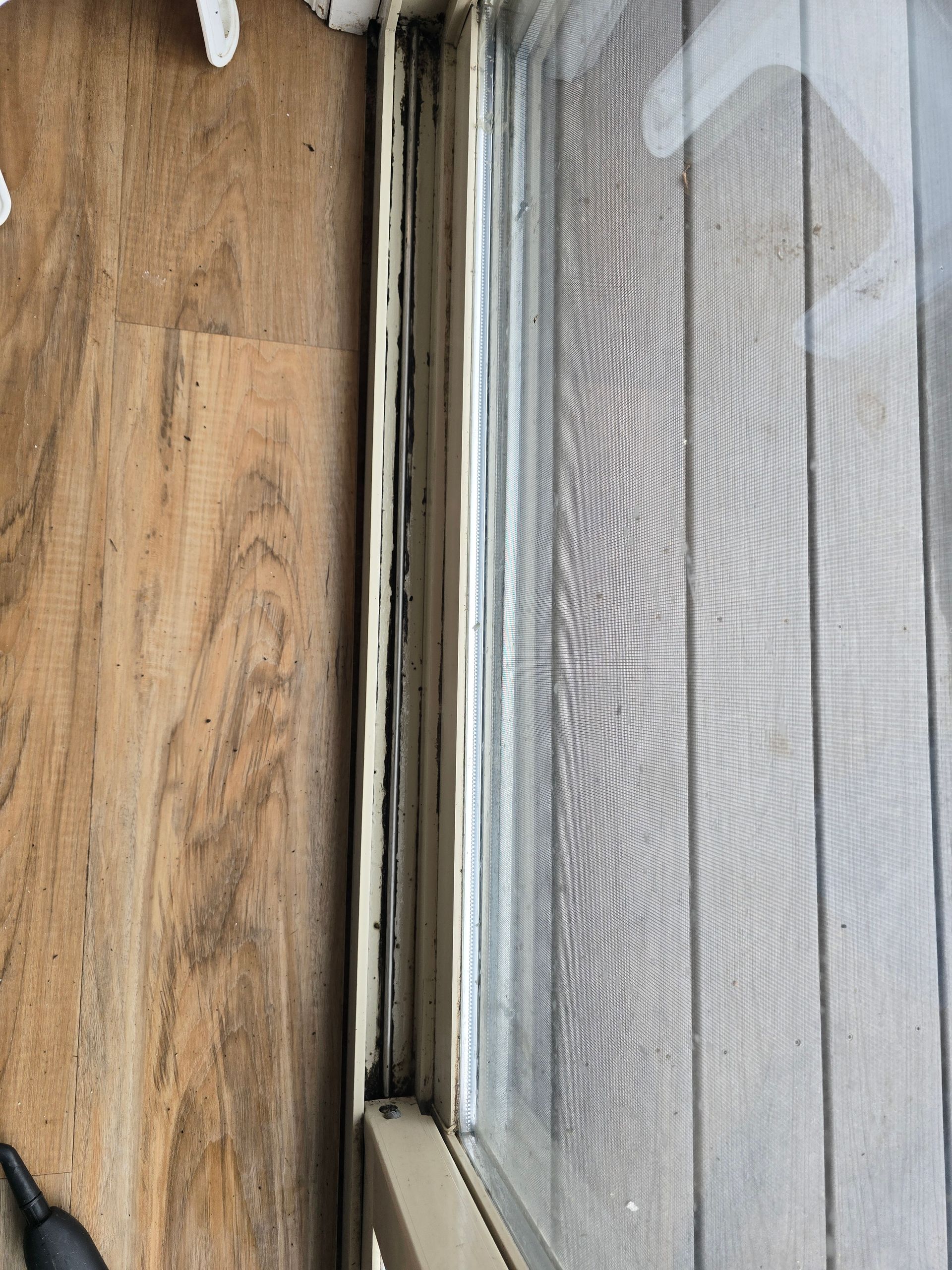 A close-up view of a worn, cream-colored window track sitting next to wood-look flooring and a window screen.