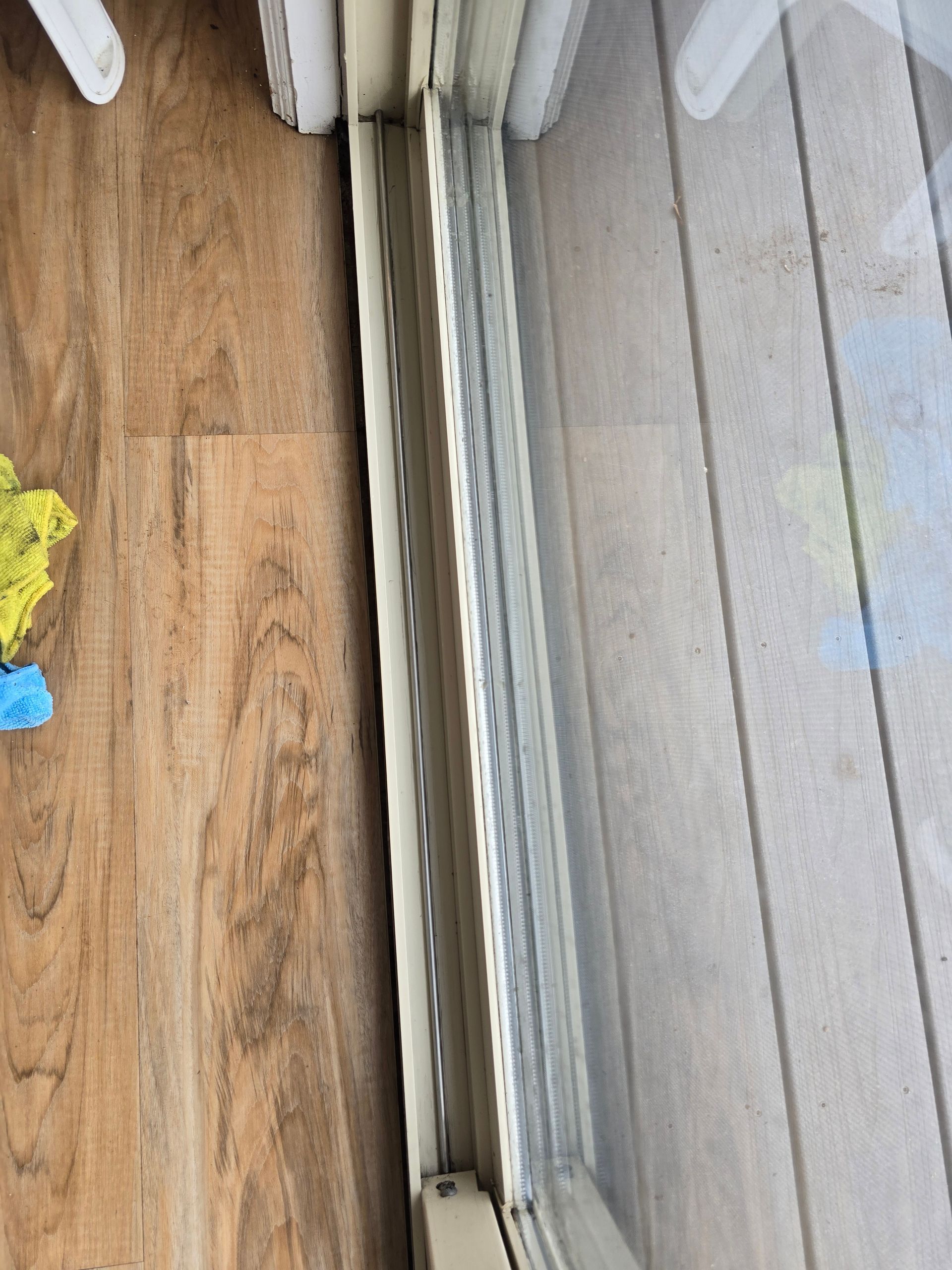 A close-up, top-down view of a sliding glass door track showing white frames, a metal rail, and adjacent wood-look flooring.