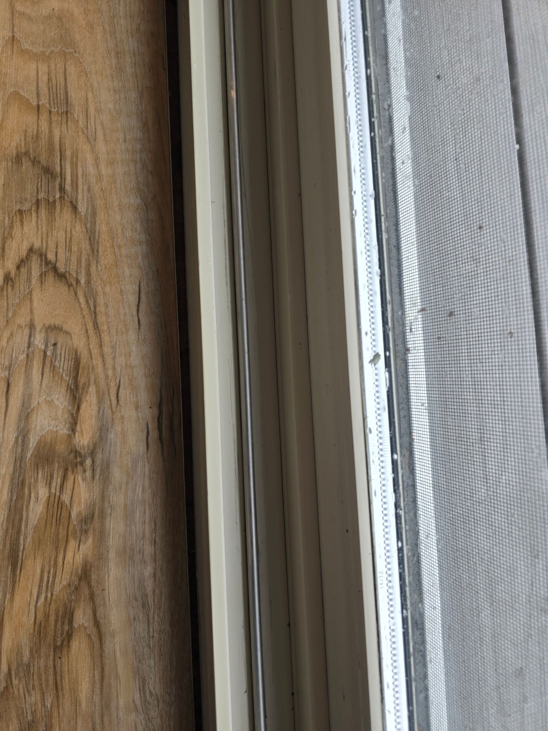 A close-up view of a vertical gap between a textured wooden wall and a white window frame with an attached mesh screen.