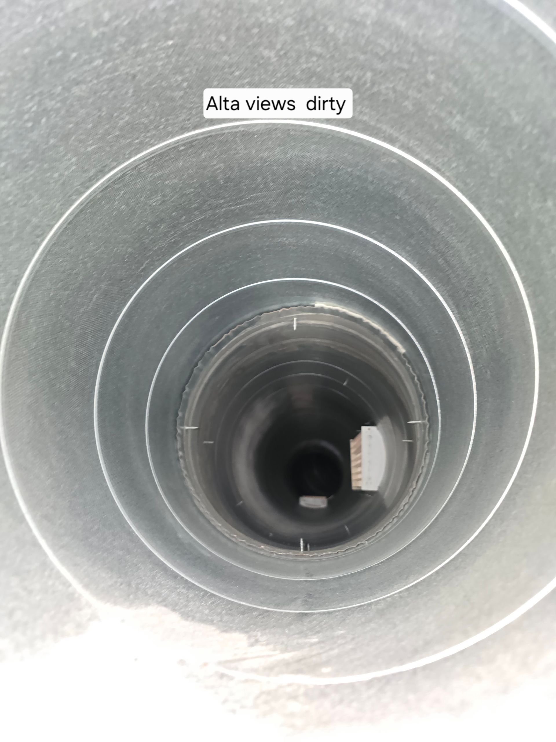 A view looking down into a cylindrical metal vent pipe with visible dirt and debris on the interior walls.