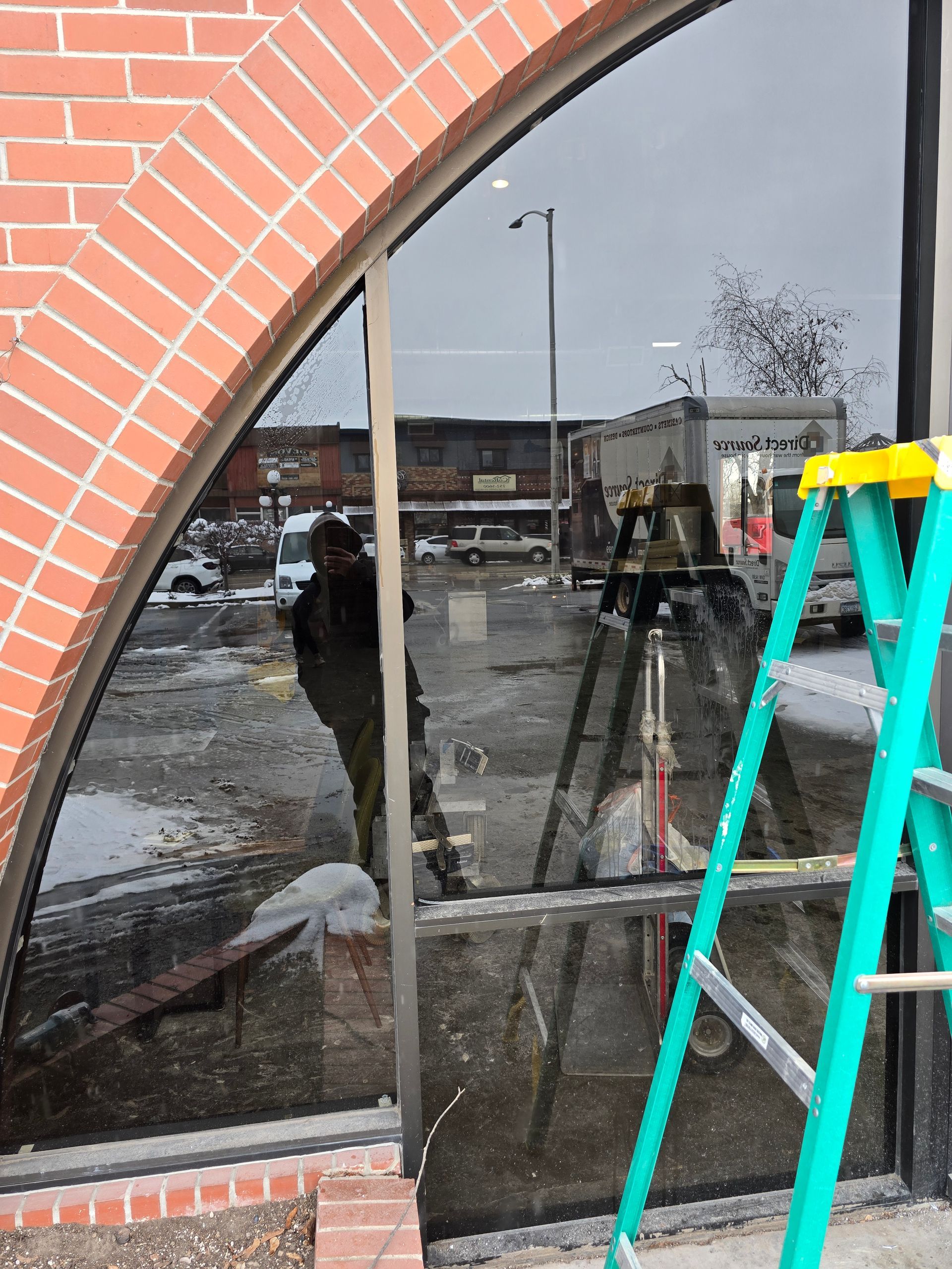 A person wearing dark clothing works on a large, arched glass storefront with a green ladder nearby.