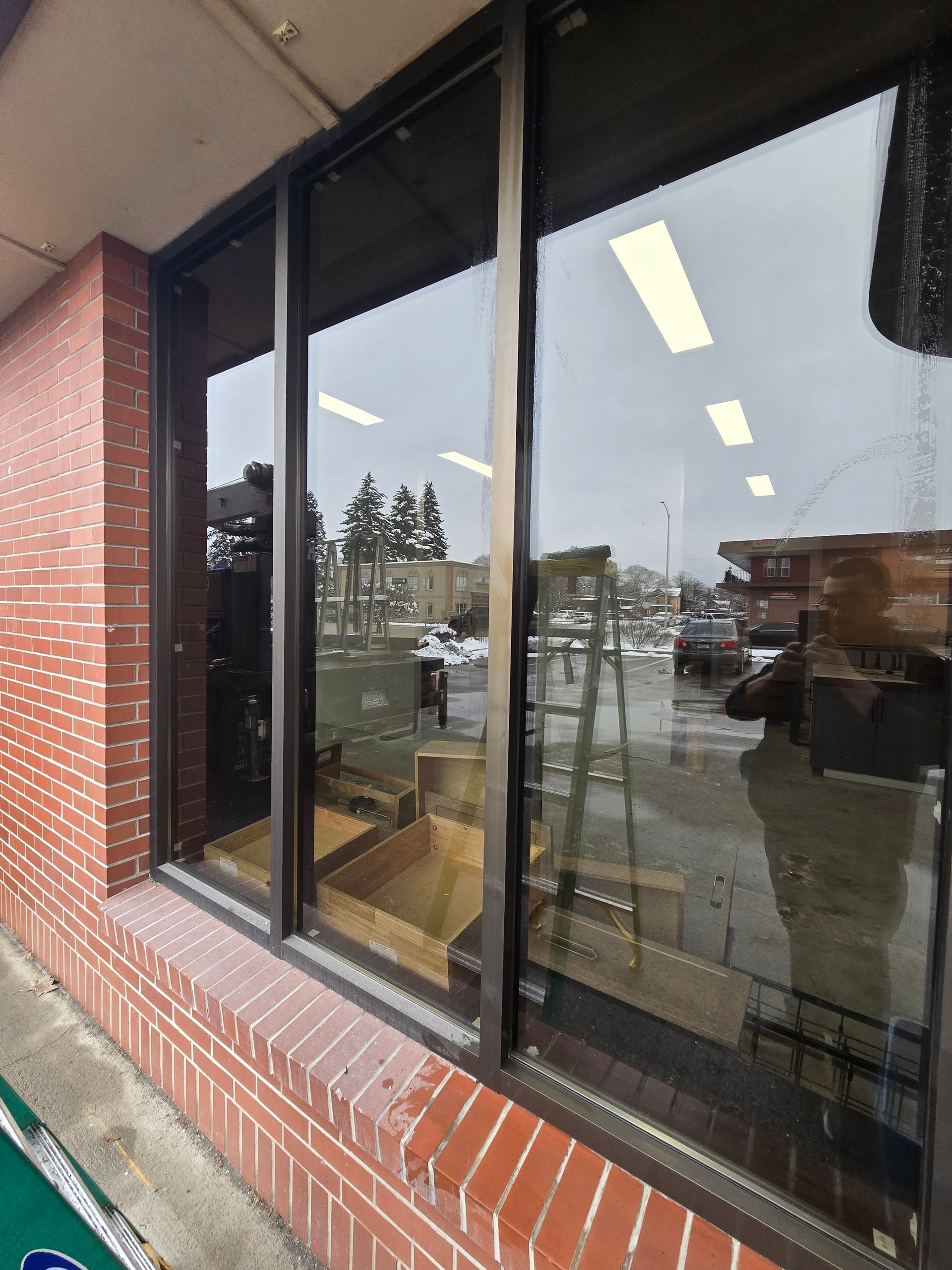 A brick building exterior featuring large glass windows reflecting a parking lot, trees, and a person holding a camera.