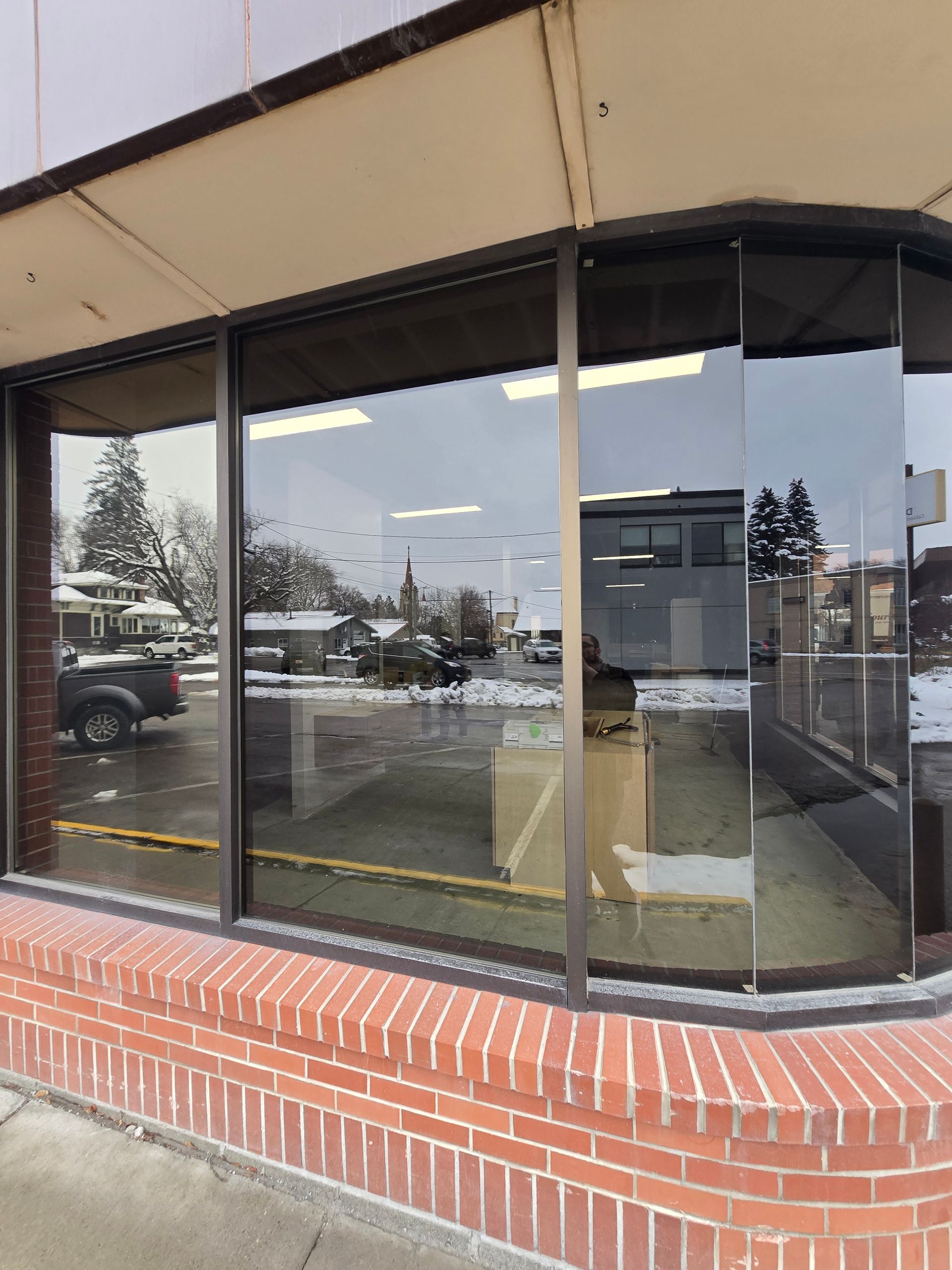 The corner of a brick-facade storefront with large glass windows reflecting a parking lot and a dark pickup truck.