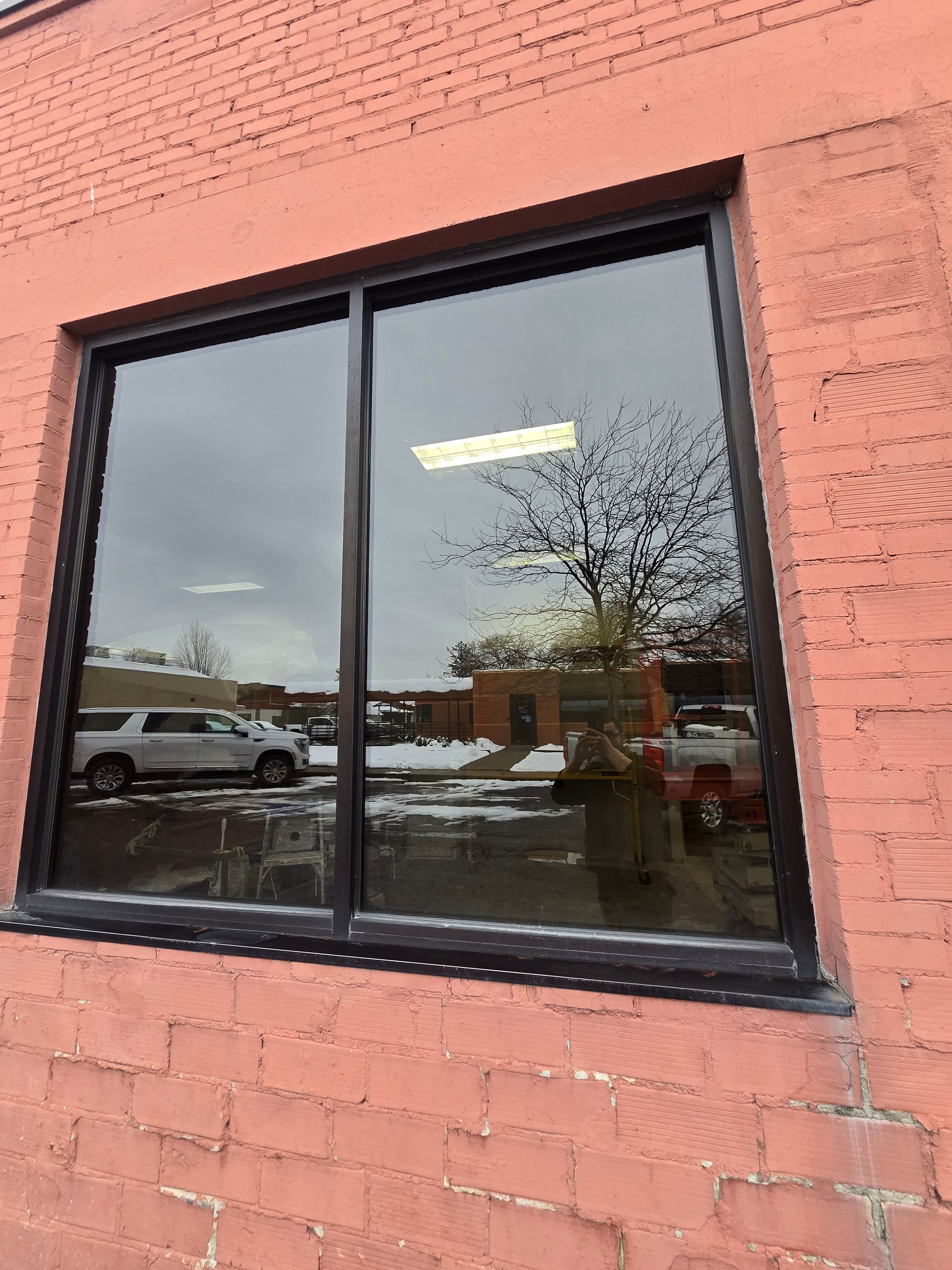 A rectangular window with a black frame installed in a textured, salmon-colored brick wall reflecting an outdoor scene.
