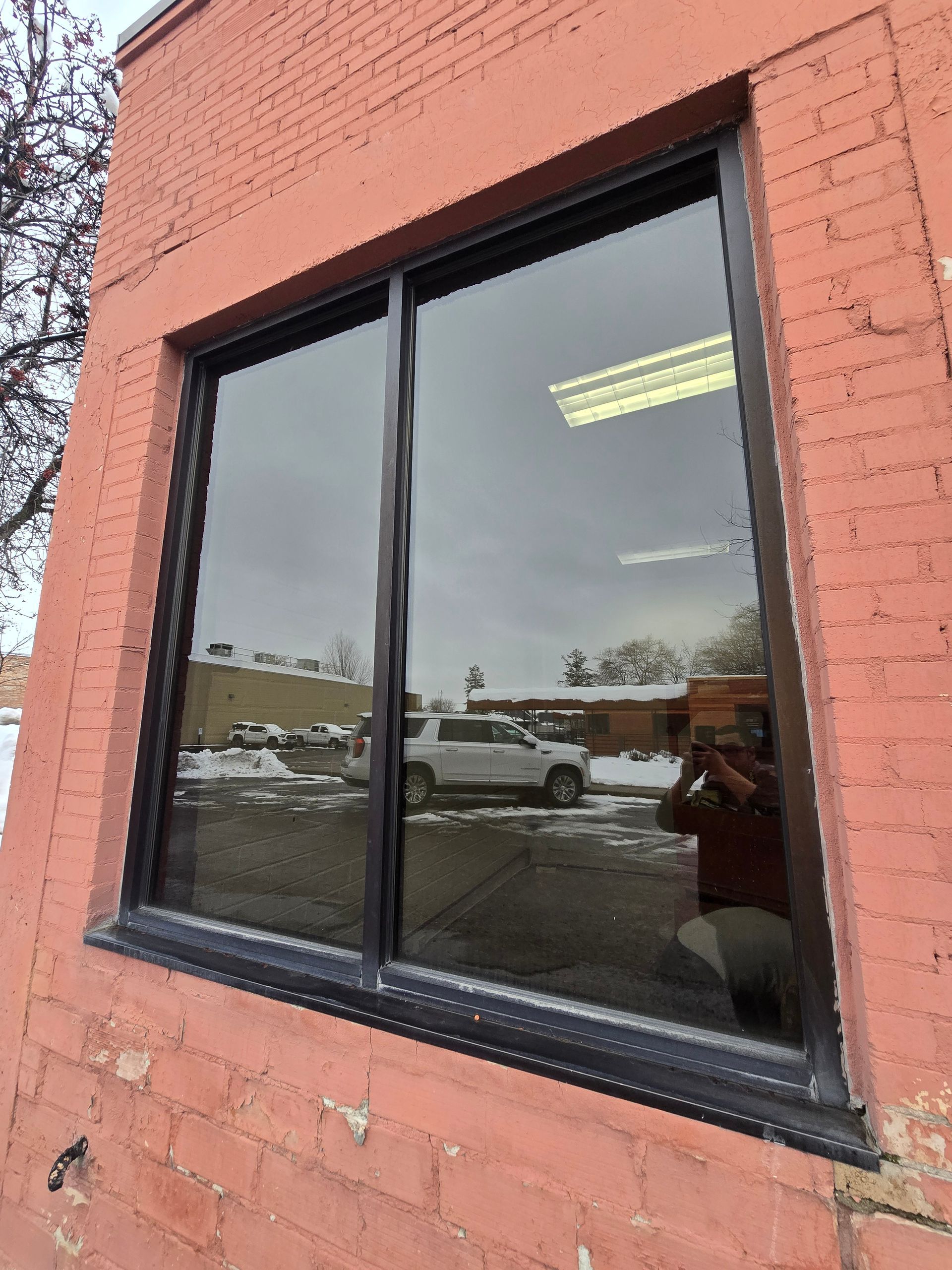 A rectangular, black-framed window on a red brick building reflecting a snowy parking lot and a parked vehicle.