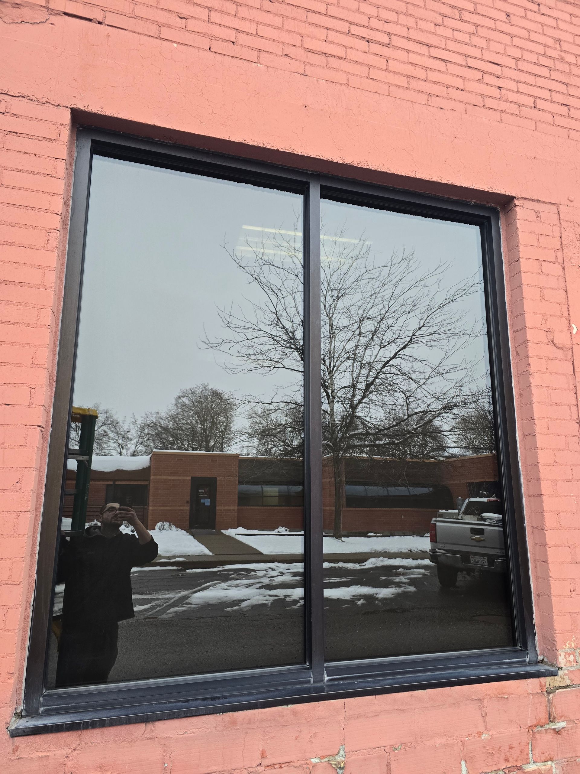 A black-framed window installed in a pink brick wall, reflecting a snowy street, trees, a building, and a vehicle.